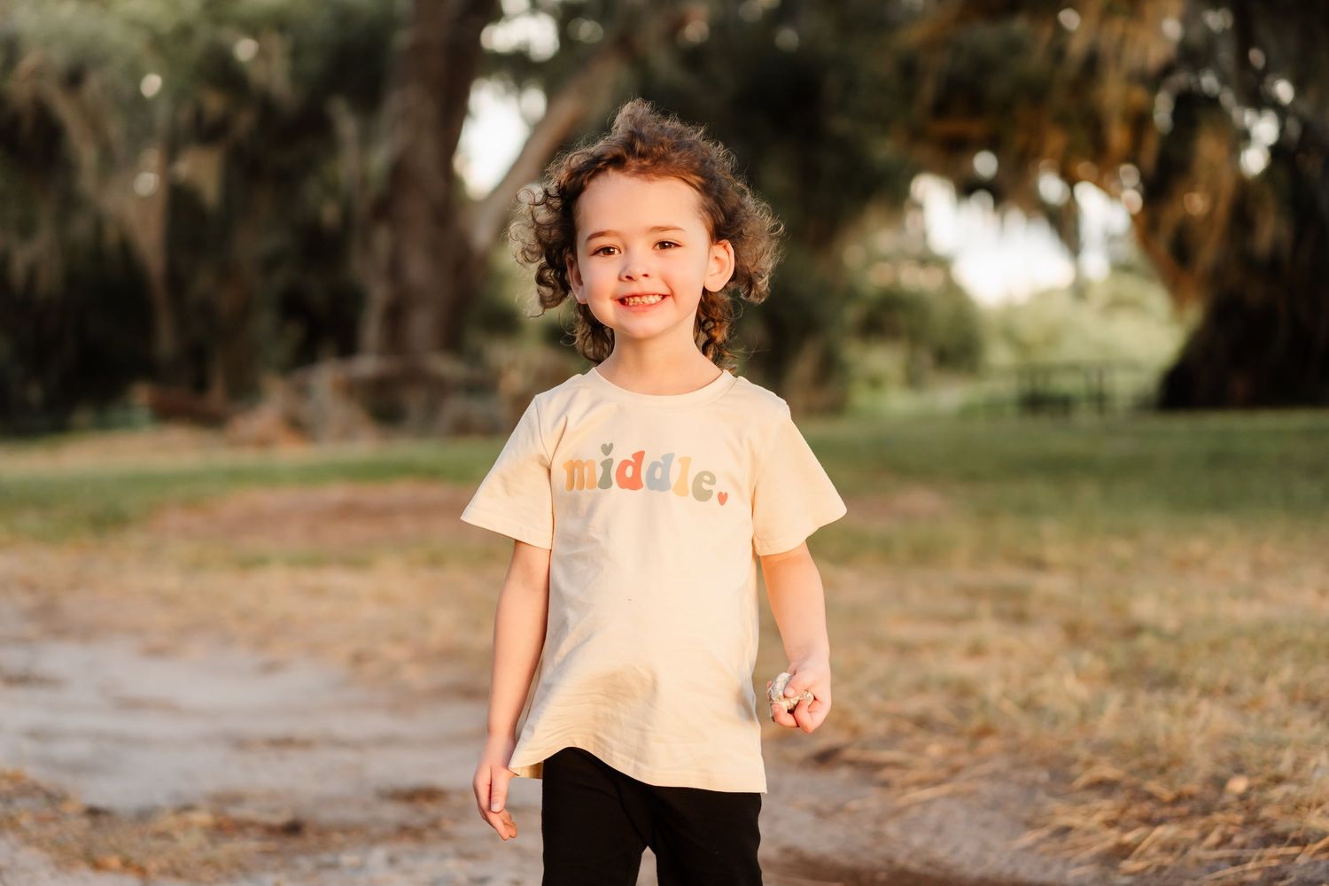 Smiling toddler wearing a cream 'middle' graphic tee and black pants standing outdoors among trees.