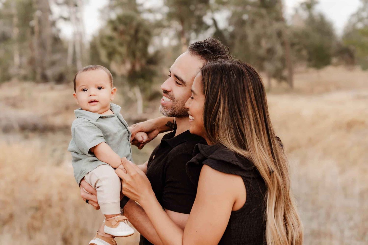 A smiling couple holds their adorable baby outdoors in a golden field surrounded by trees.