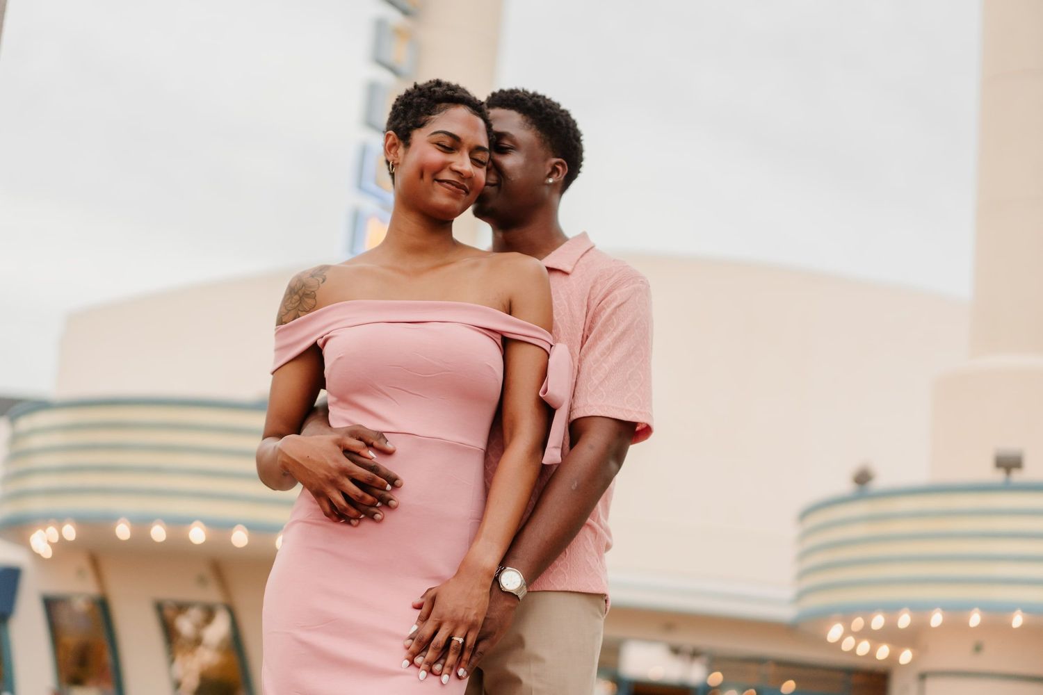 A couple shares a romantic moment, man kissing woman's cheek, both dressed in pink outfits outdoors.