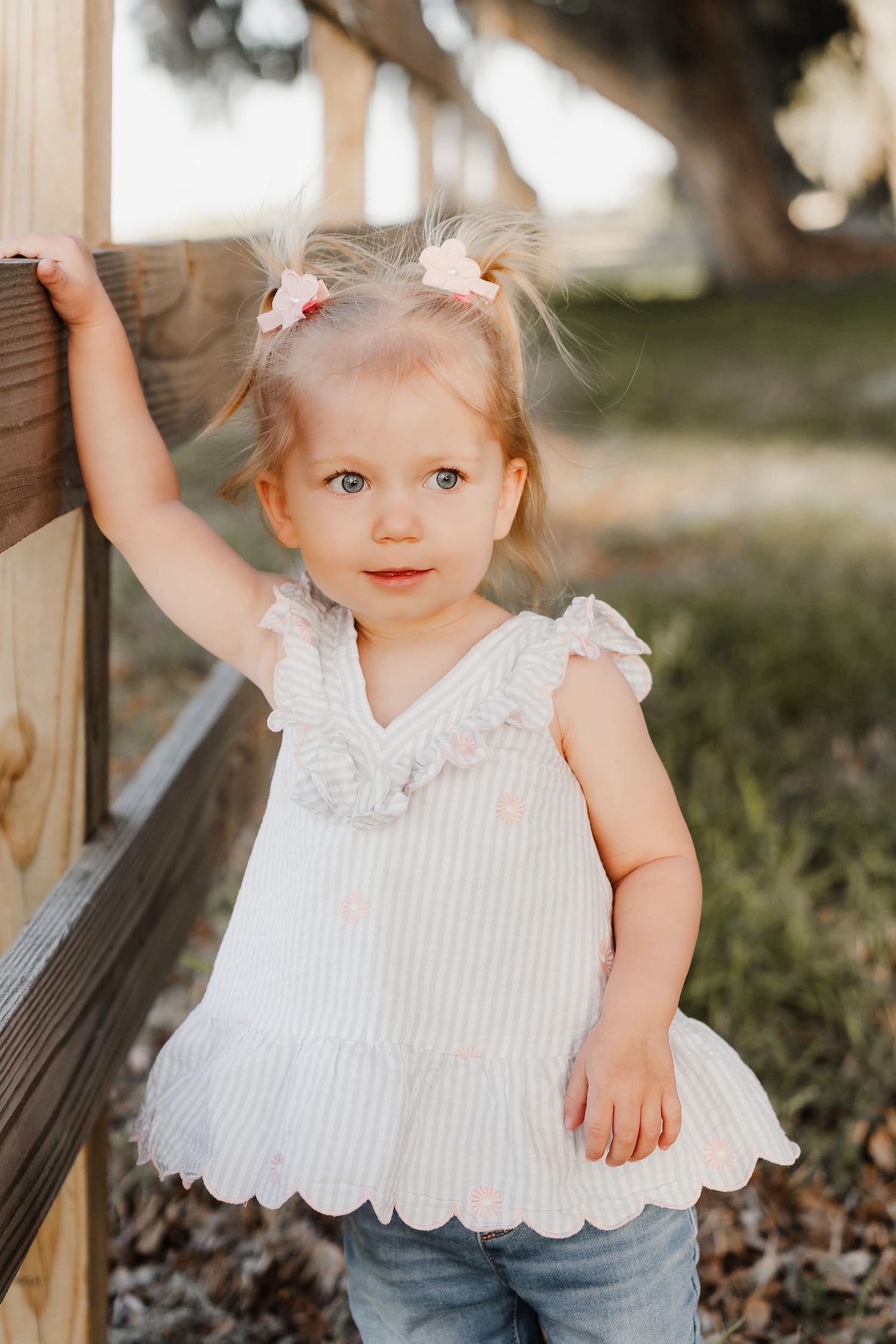 Adorable toddler girl in white ruffle top and jeans stands by wooden fence outdoors in warm sunlight.