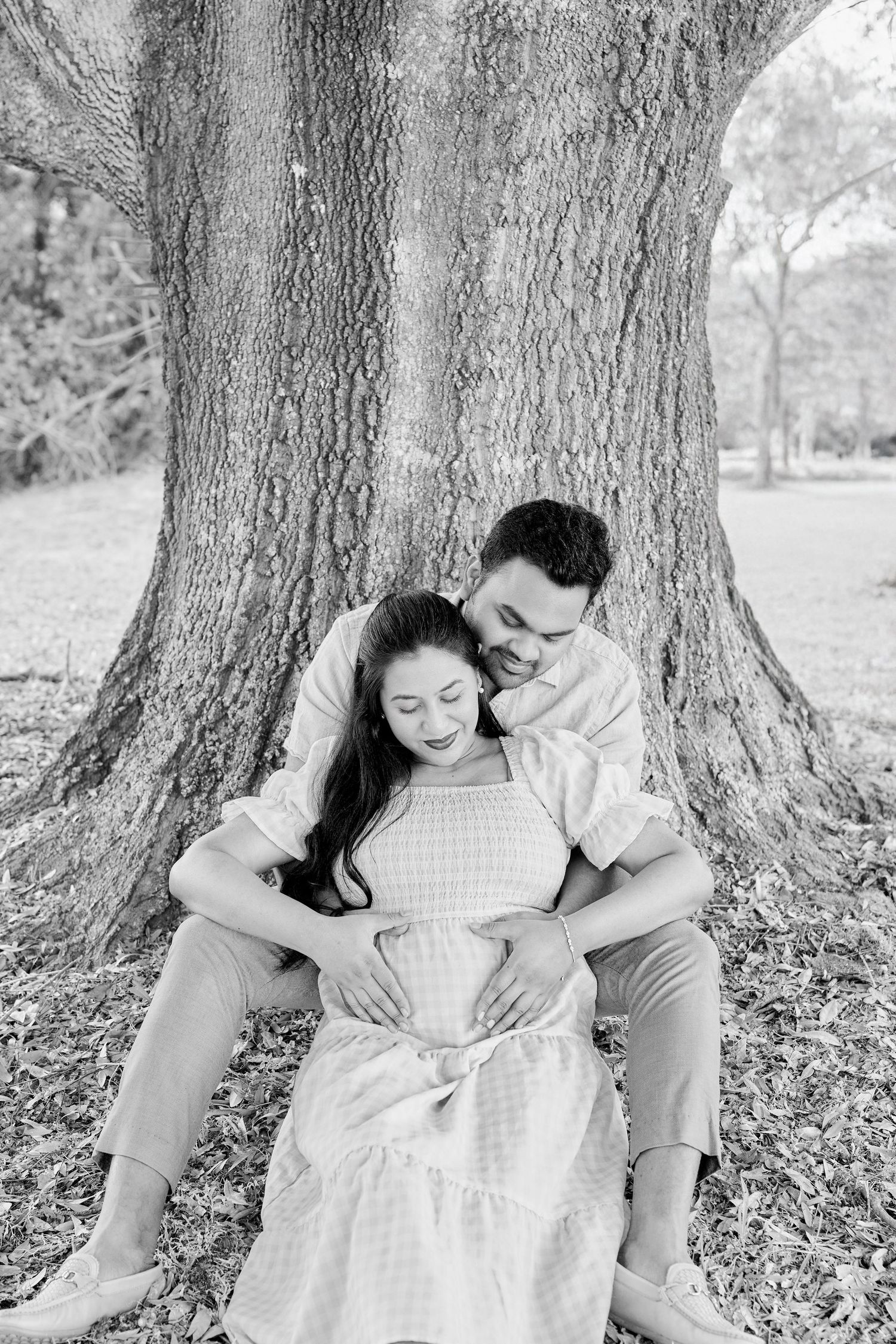 Black and white photo of a couple sitting against a large tree, embracing tenderly during a maternity photoshoot.