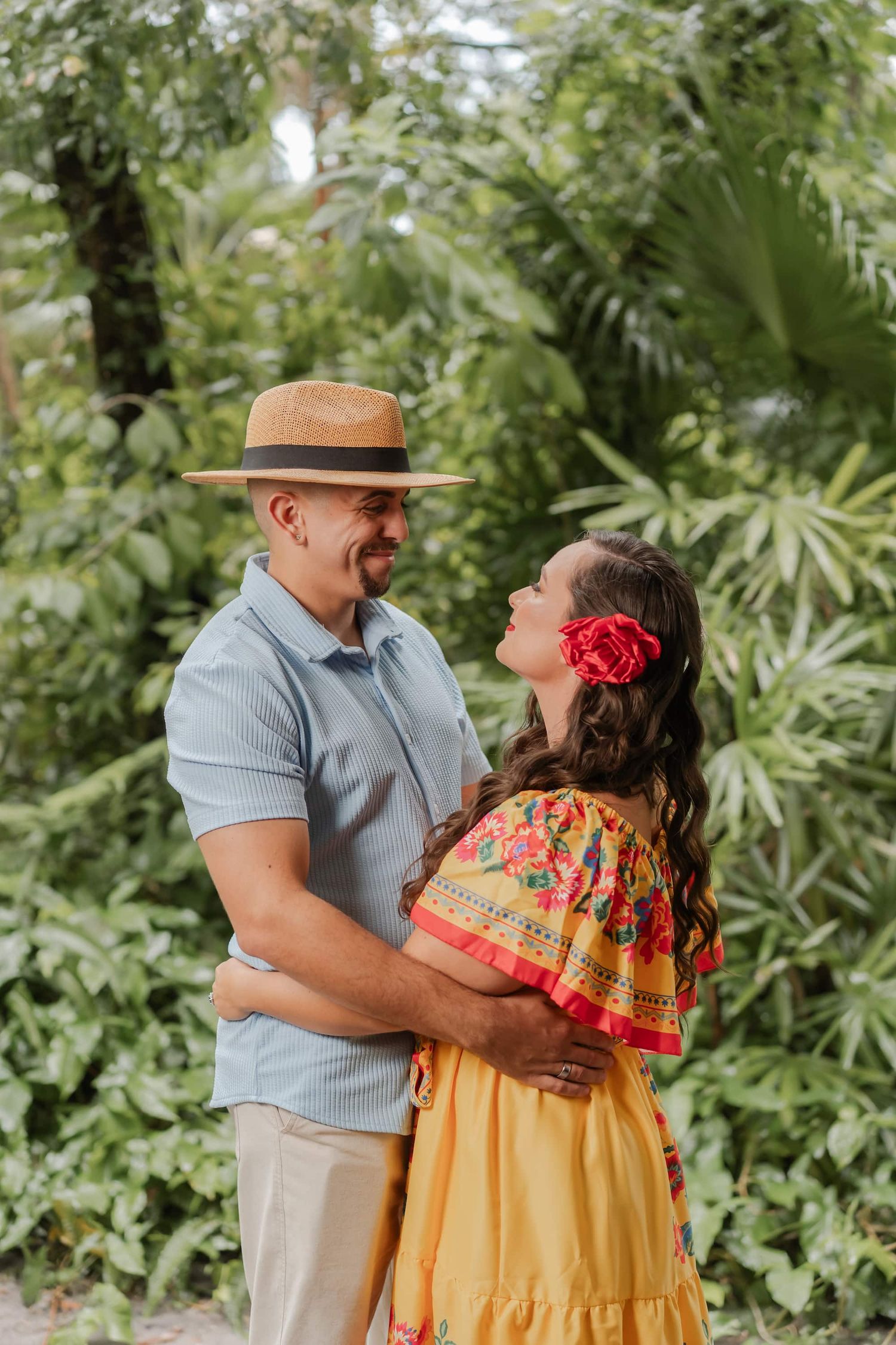 Smiling couple embracing in a lush tropical garden, he wears a straw hat and she a colorful embroidered yellow dress with a red flower.