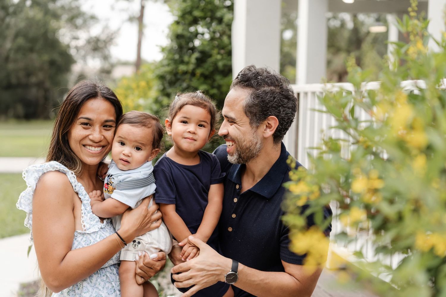 Happy family of four with two young children smiling outdoors near a white porch surrounded by greenery.