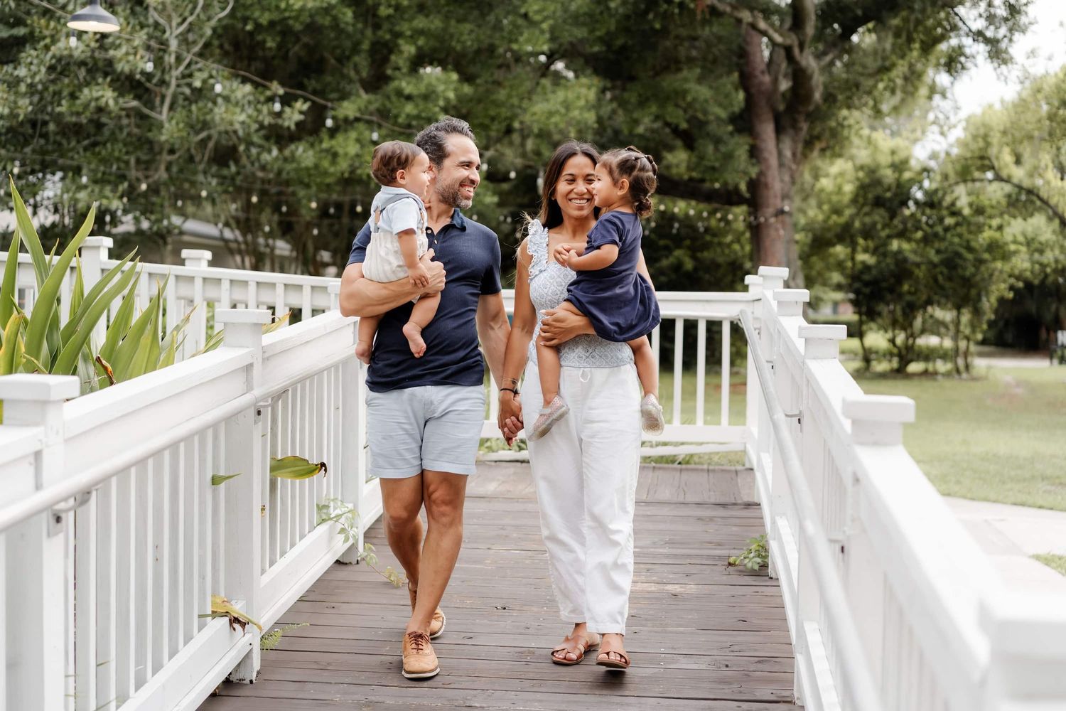 Happy family with two toddlers walking on a white wooden bridge surrounded by lush green trees.