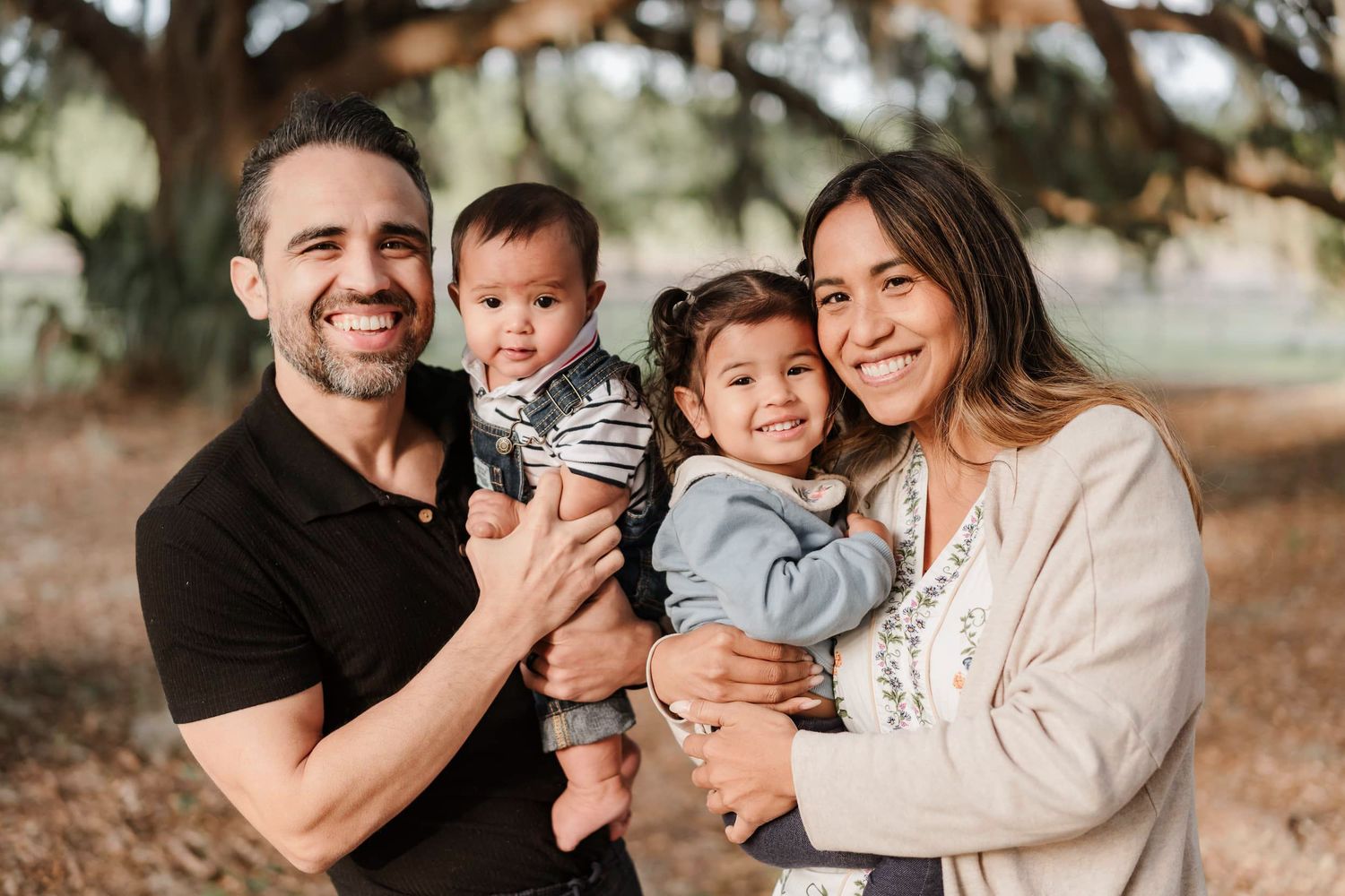 A smiling family of four poses outdoors, with parents holding two young children among trees in a warm, natural setting.