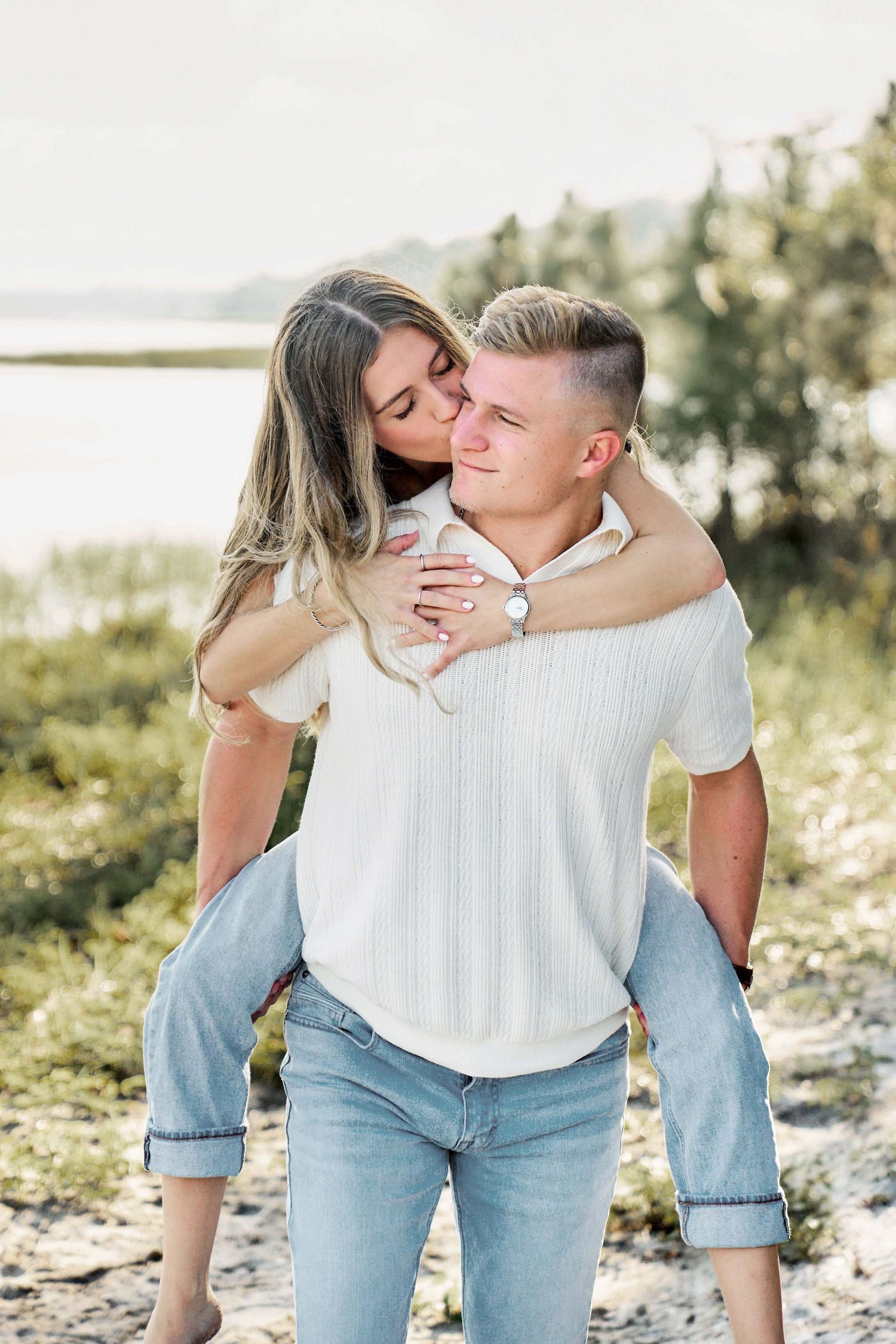 A romantic couple in casual attire share tender moments on a sunny beach with soft natural lighting.