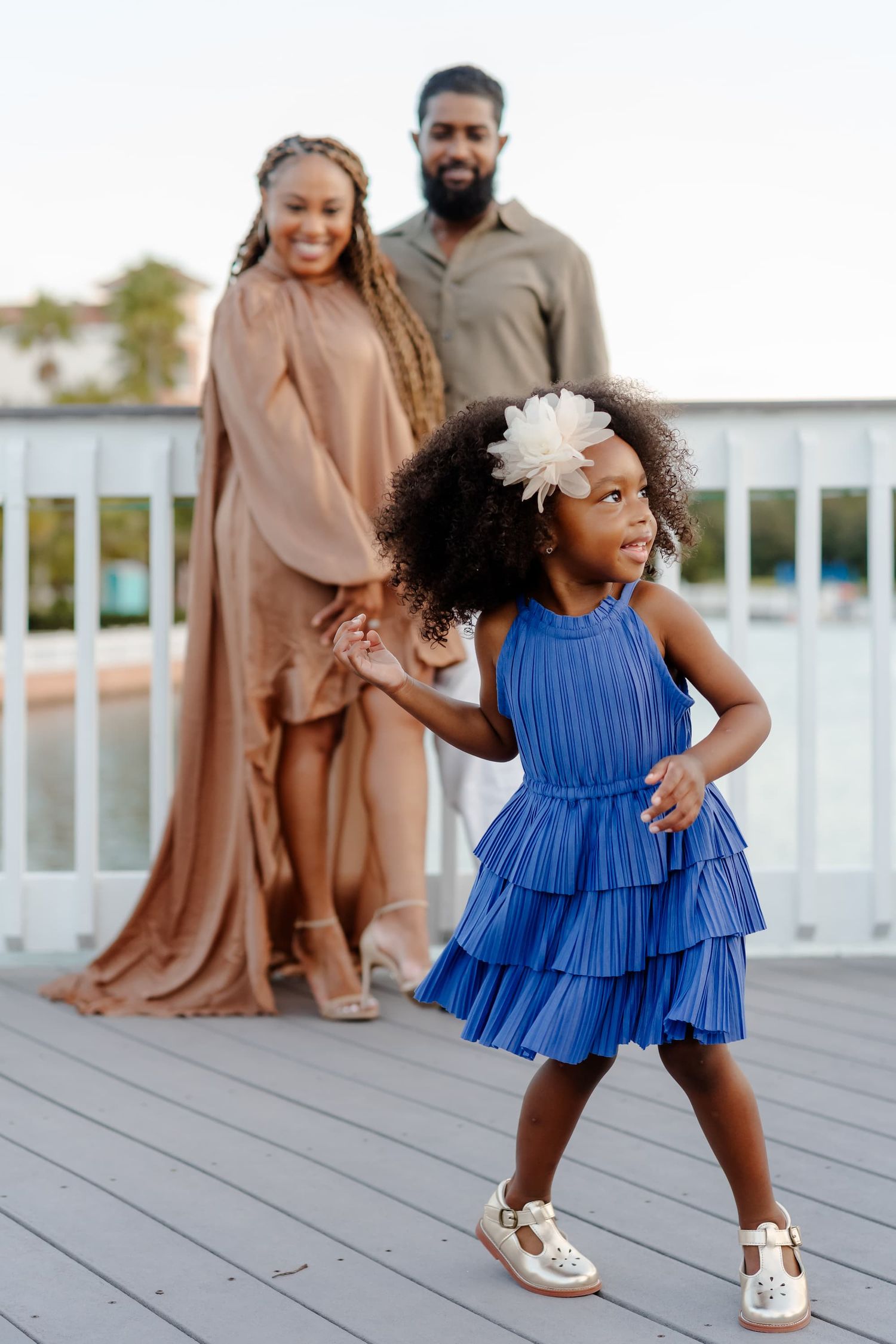 A young girl in a blue tiered dress and white flower headband twirls on a wooden deck while smiling parents stand behind her.