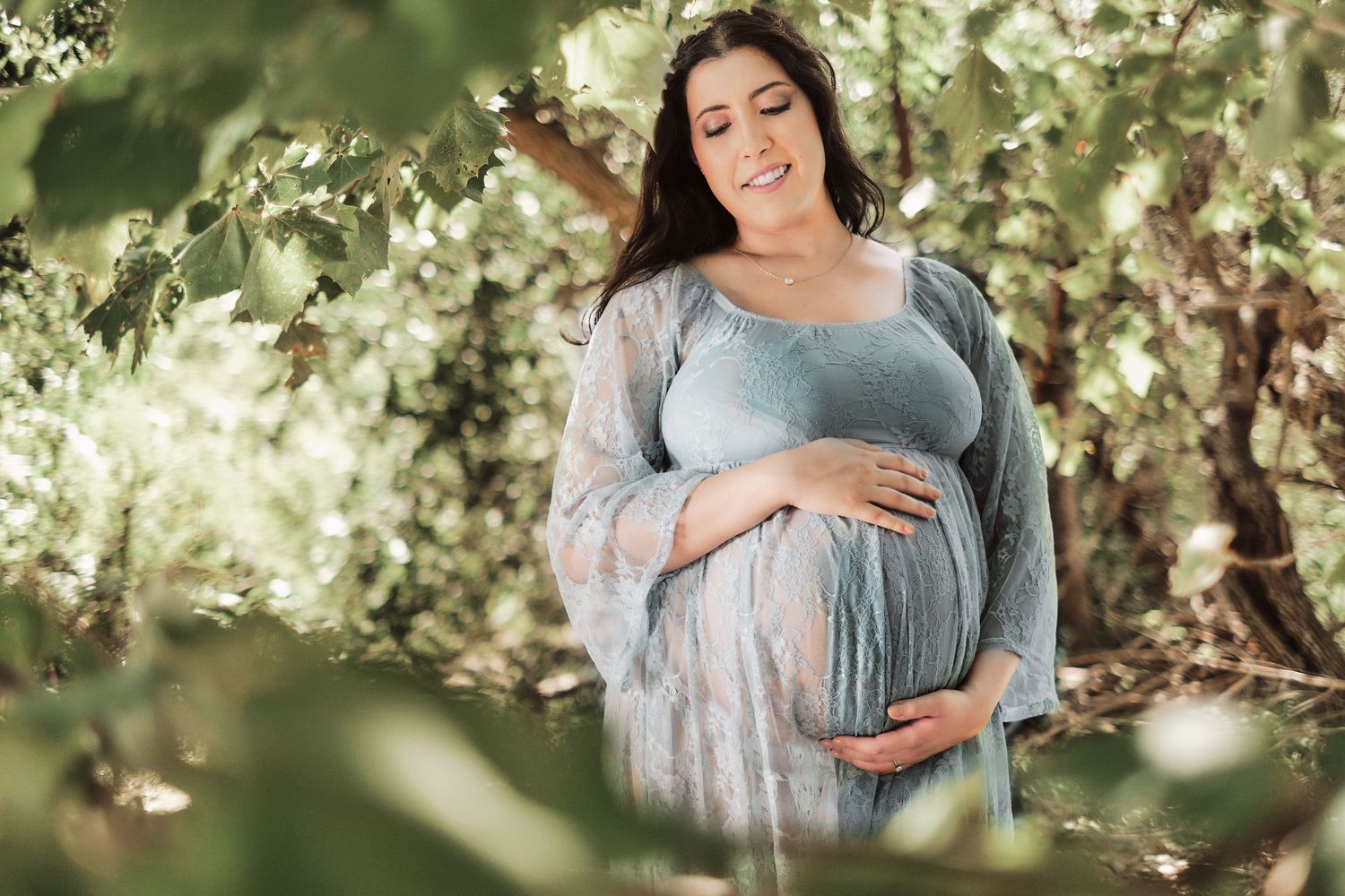 Pregnant woman in blue dress cradling baby bump outdoors among green trees during maternity photo session.