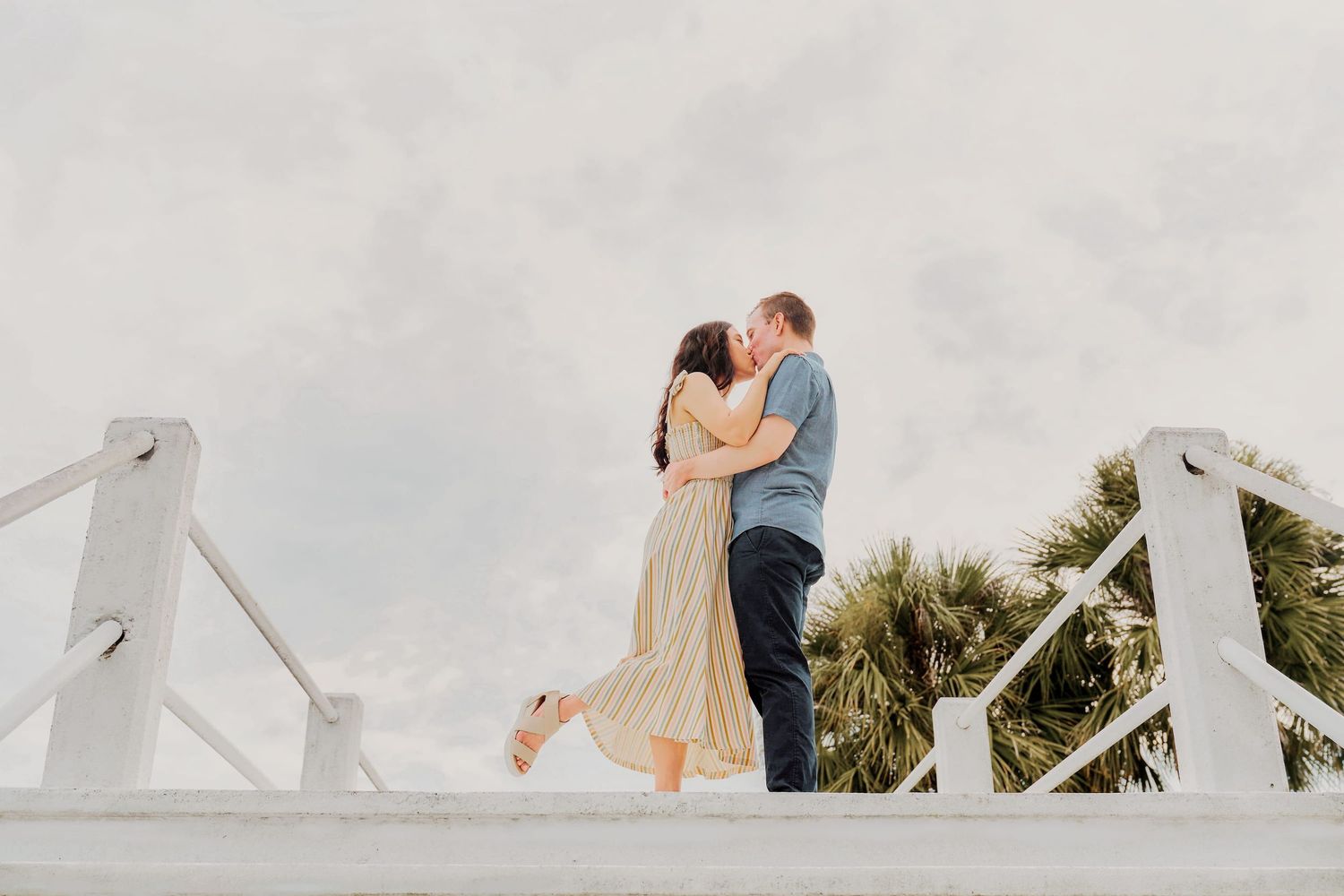 Couple sharing a romantic kiss on a white bridge railing with palm trees and cloudy sky backdrop.