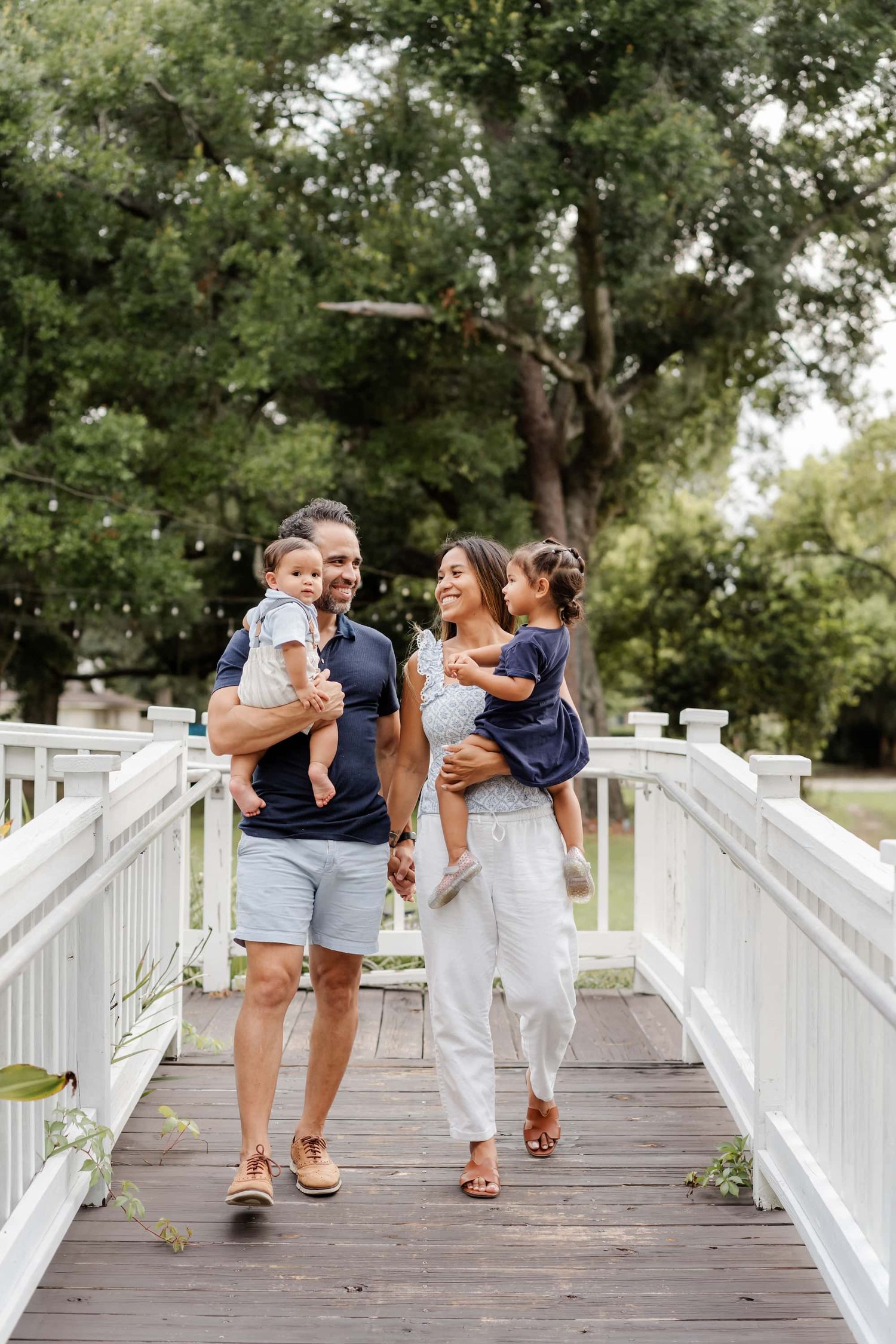 A happy family of four walks on a white wooden bridge surrounded by lush green trees.