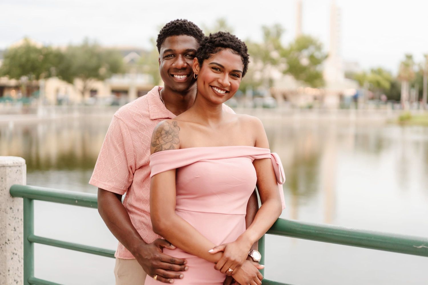 A happy Black couple in matching pink outfits embracing on a waterfront bridge, smiling warmly at the camera.