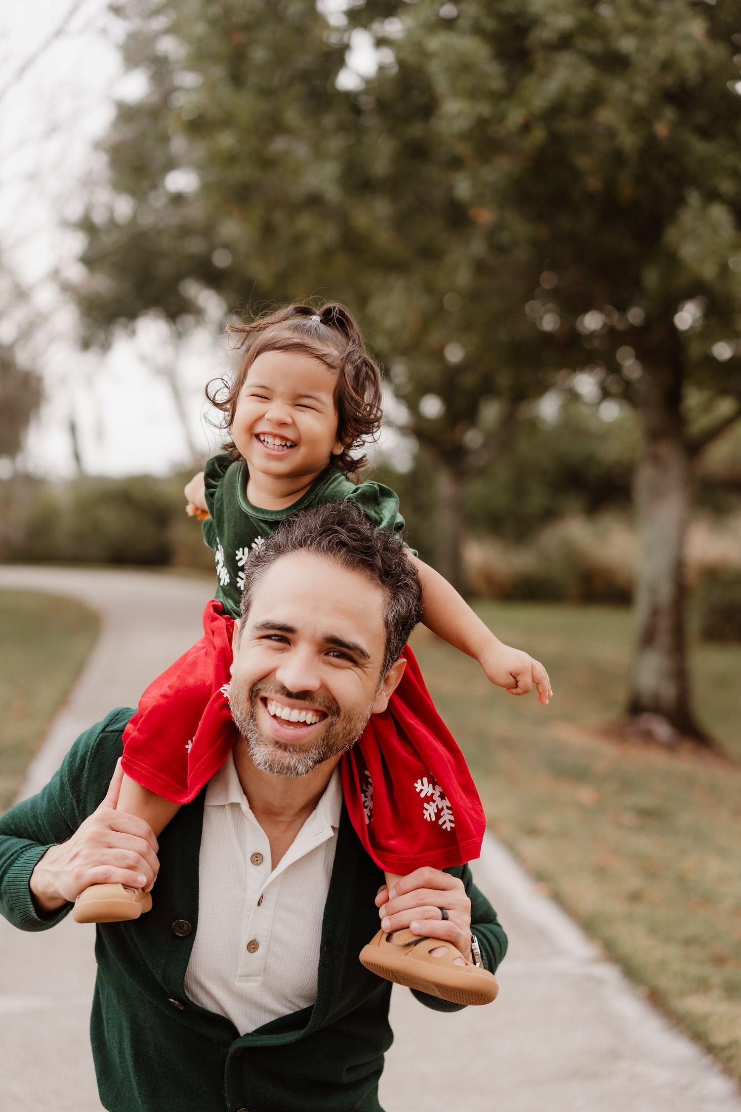 Smiling man carrying a laughing young girl on his shoulders outdoors in a park setting, both wearing festive green and red.