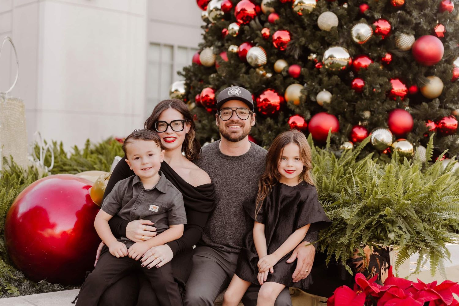 Family of four posing in front of decorated Christmas tree with red ornaments and poinsettias.