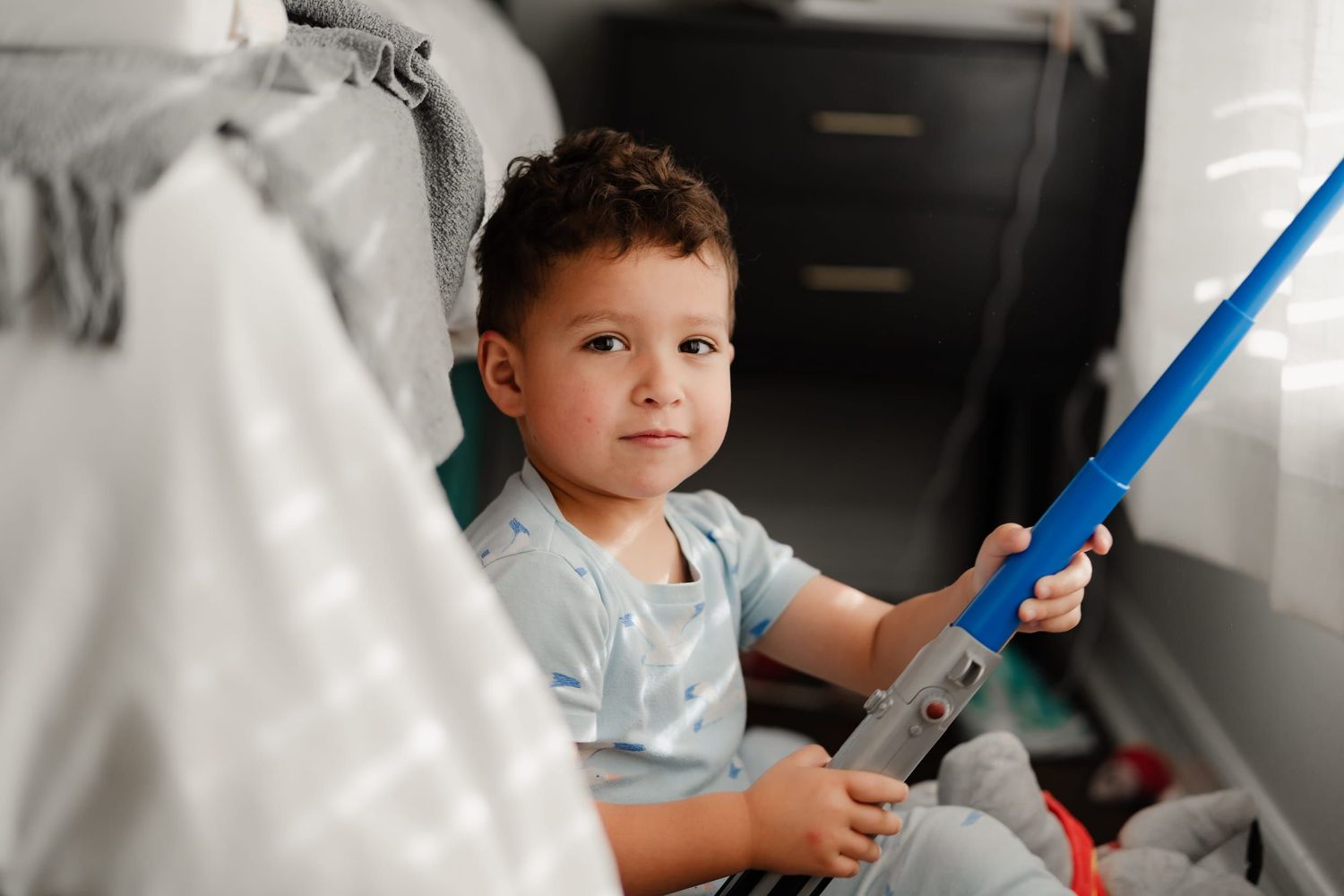 A young toddler boy sits in an airplane seat, holding a blue seatbelt, wearing a light blue shirt.