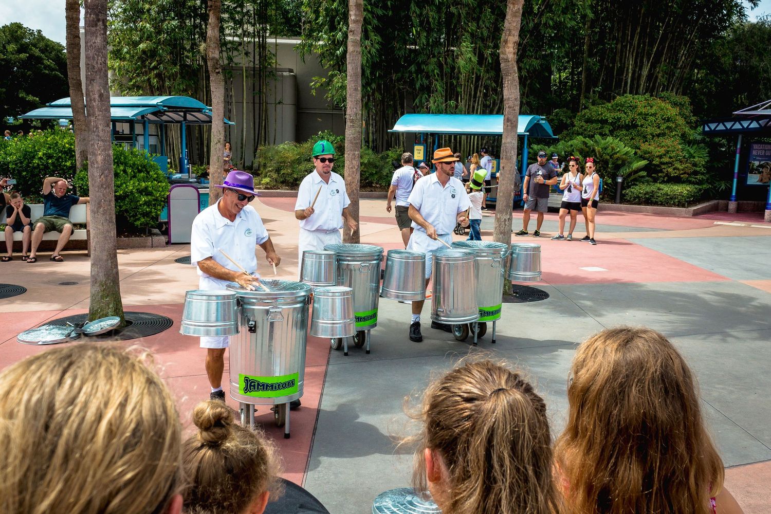 Steel drum band performs outdoors at a theme park while an audience watches on a sunny day.