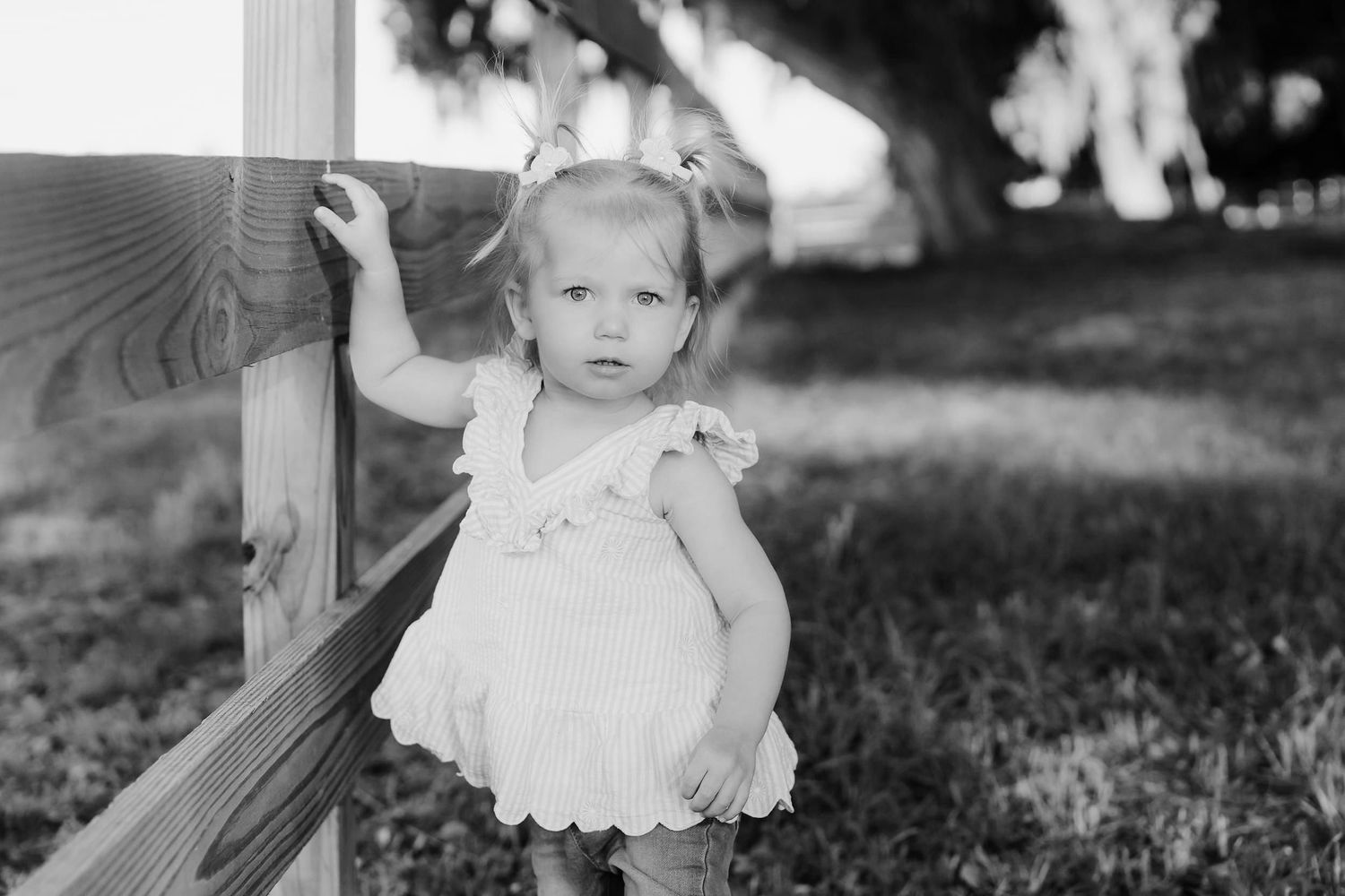 Black and white photo of a cute toddler girl in a white dress leaning against a wooden fence outdoors.