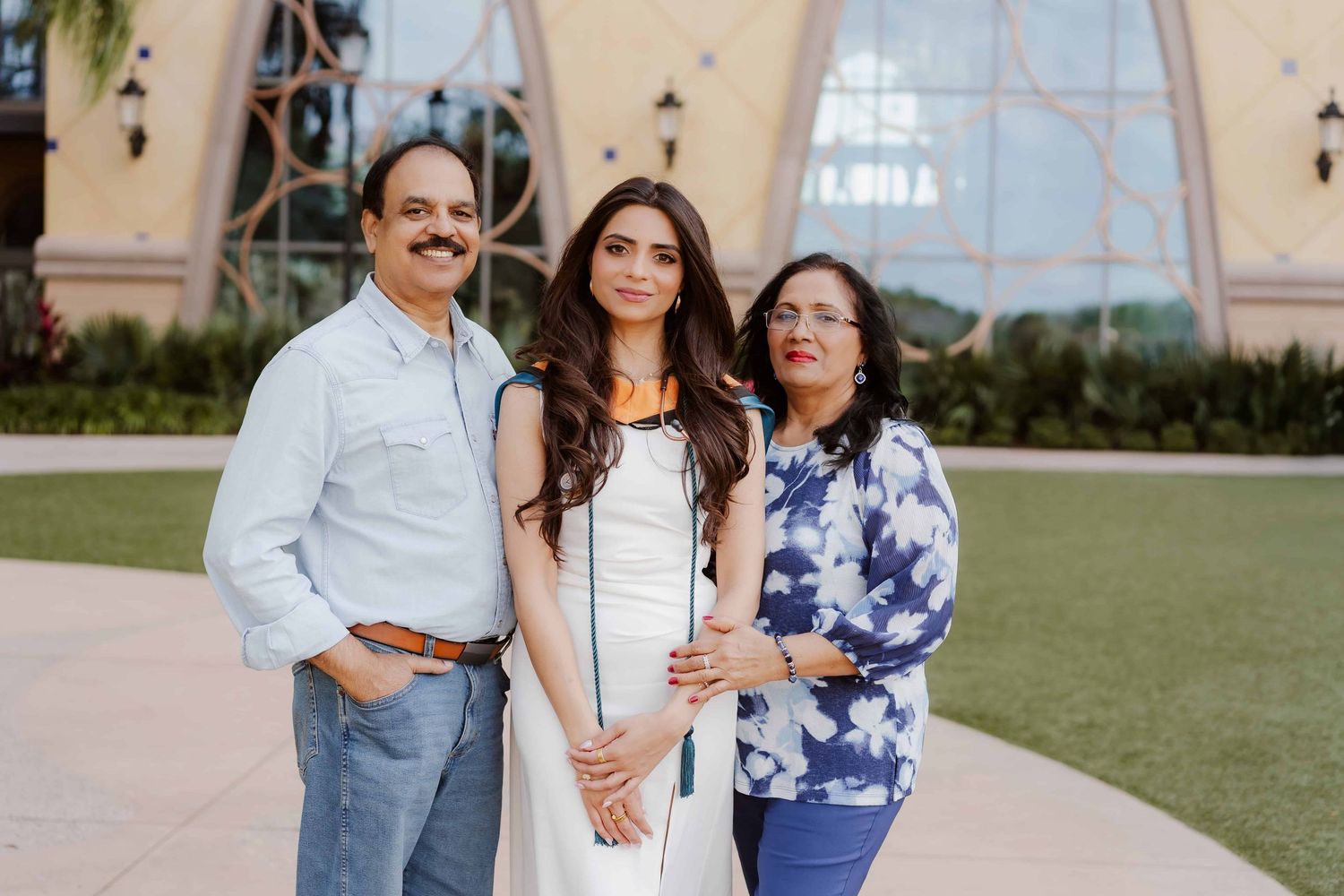 Three people stand together smiling on a grassy lawn with a pink architectural structure in the background.