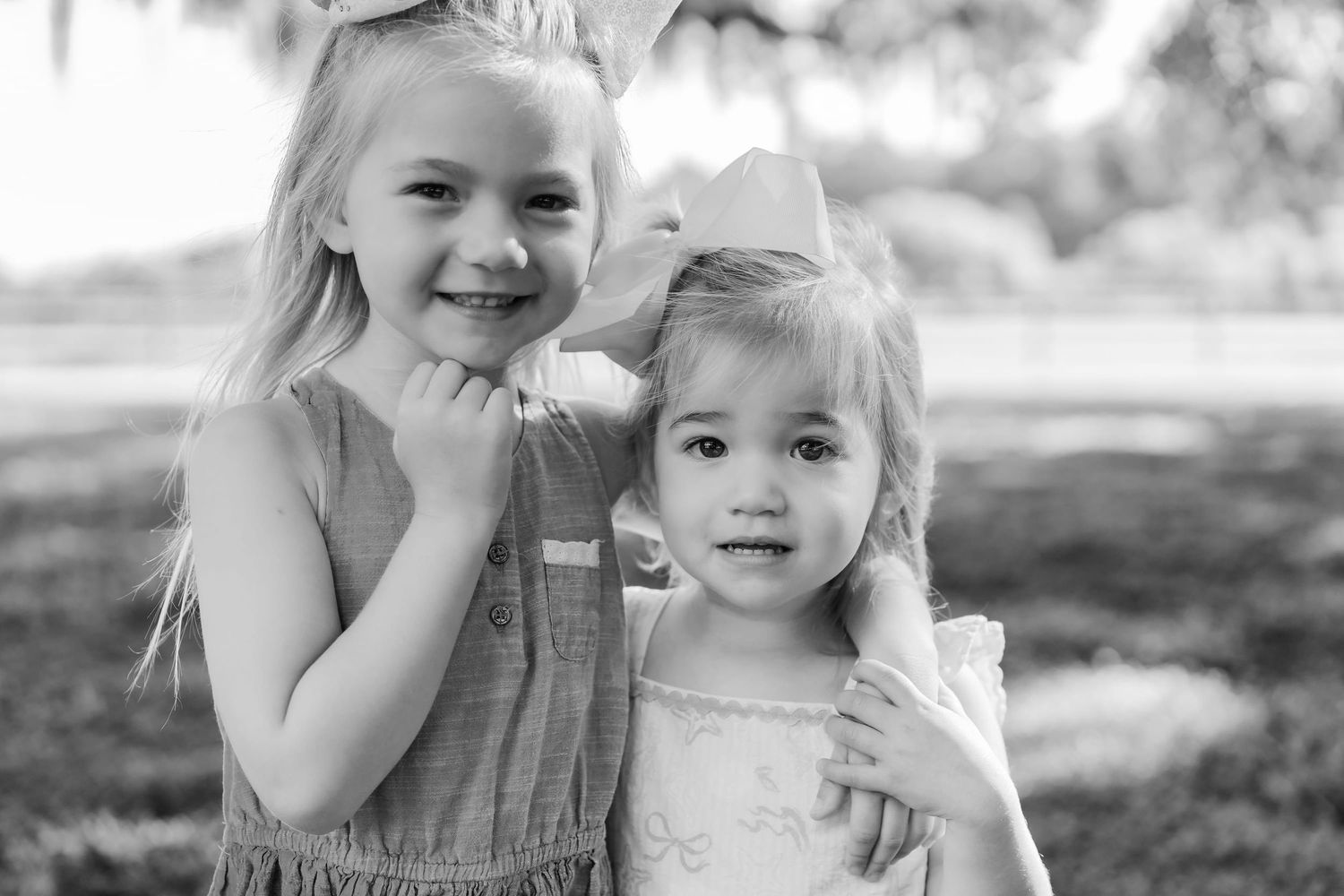 Two young girls pose outdoors in a black and white photo, the older girl hugging the younger one warmly.