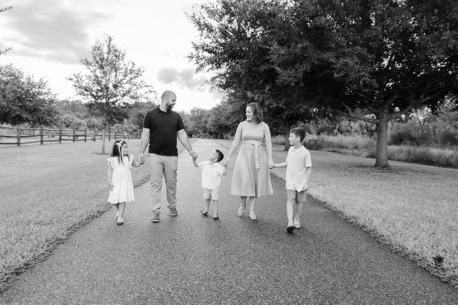 Black and white photo of a family of five walking hand-in-hand down a tree-lined path outdoors.