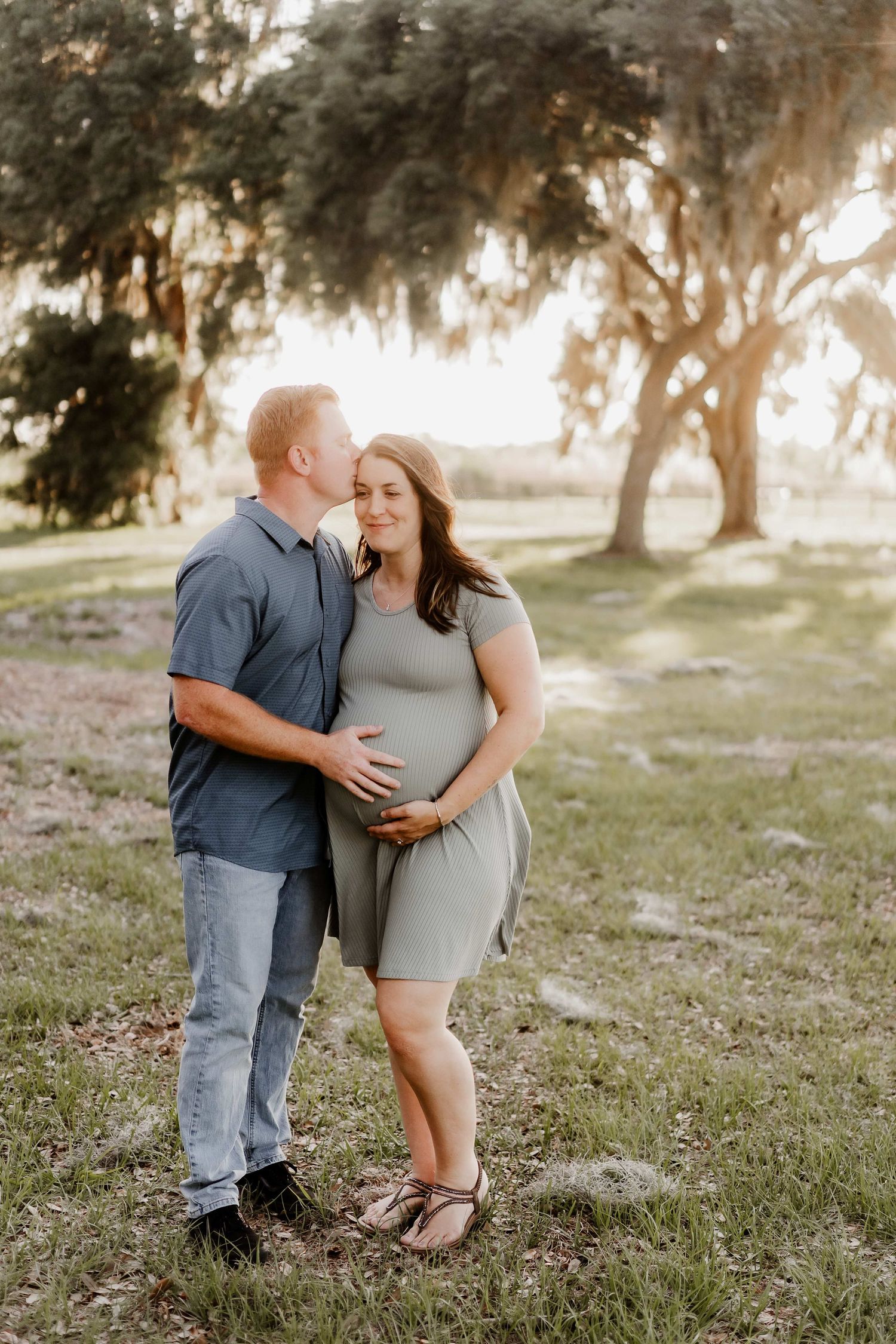 A couple shares a tender moment outdoors, with the man kissing his pregnant partner's cheek while they cradle her baby bump.