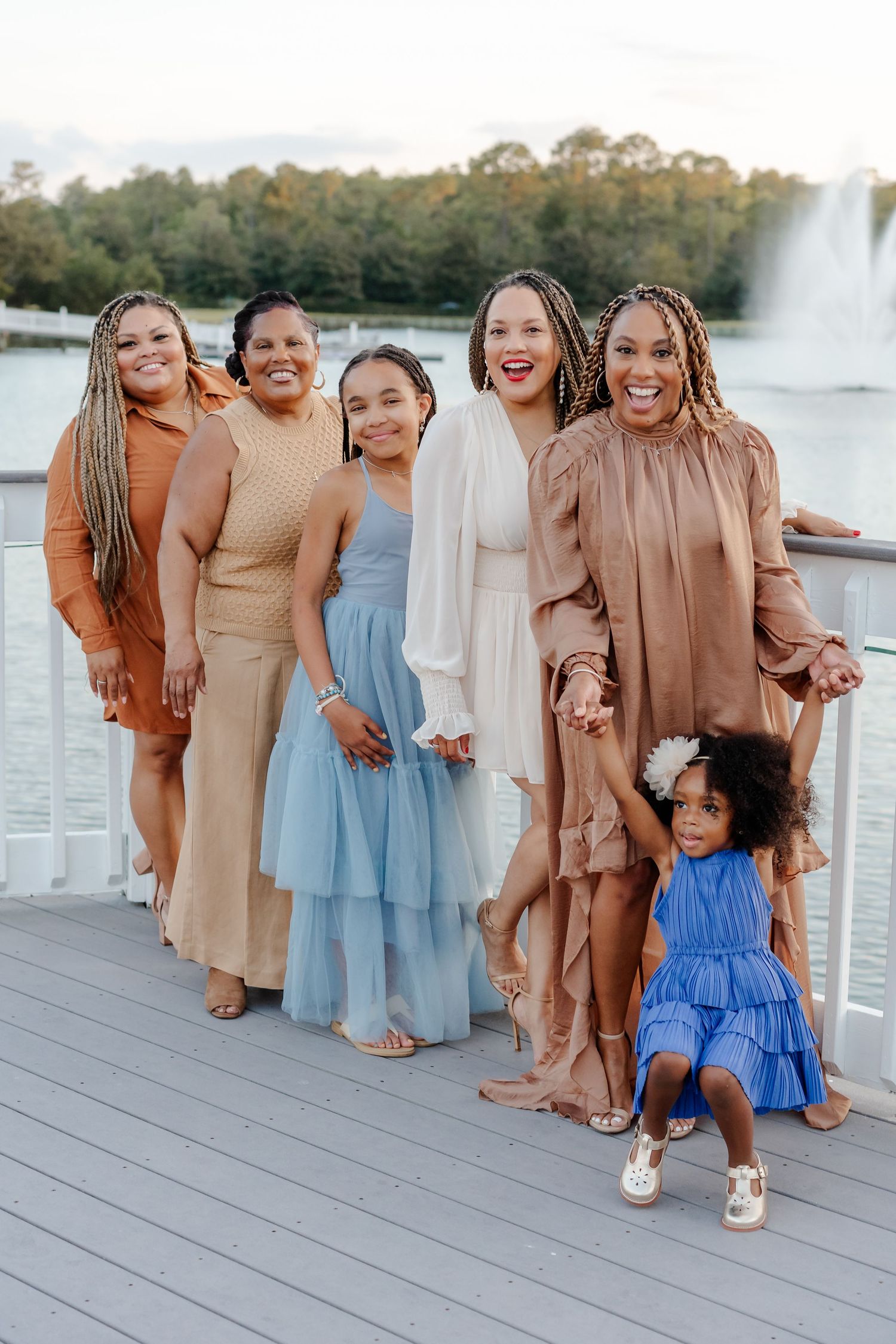 A joyful group of women and girls in elegant earth-tone and blue dresses posing on a lakeside deck.