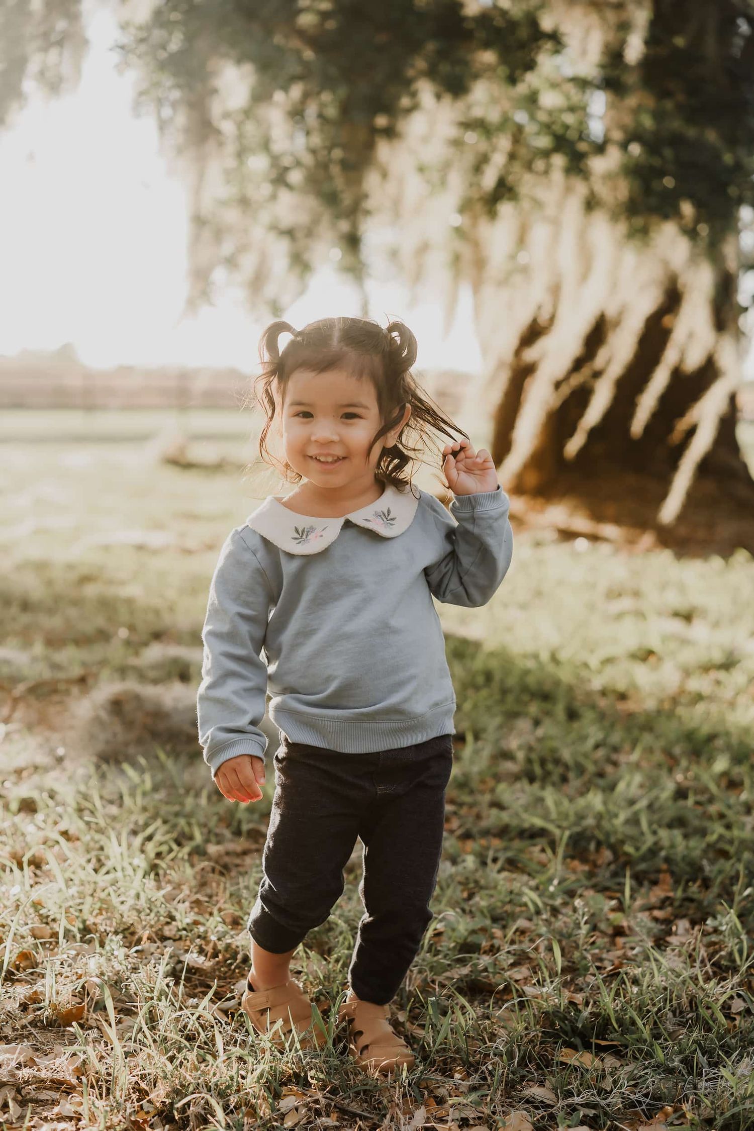 Toddler girl in blue sweatshirt with white collar and dark pants smiling outdoors near a large tree in golden light.