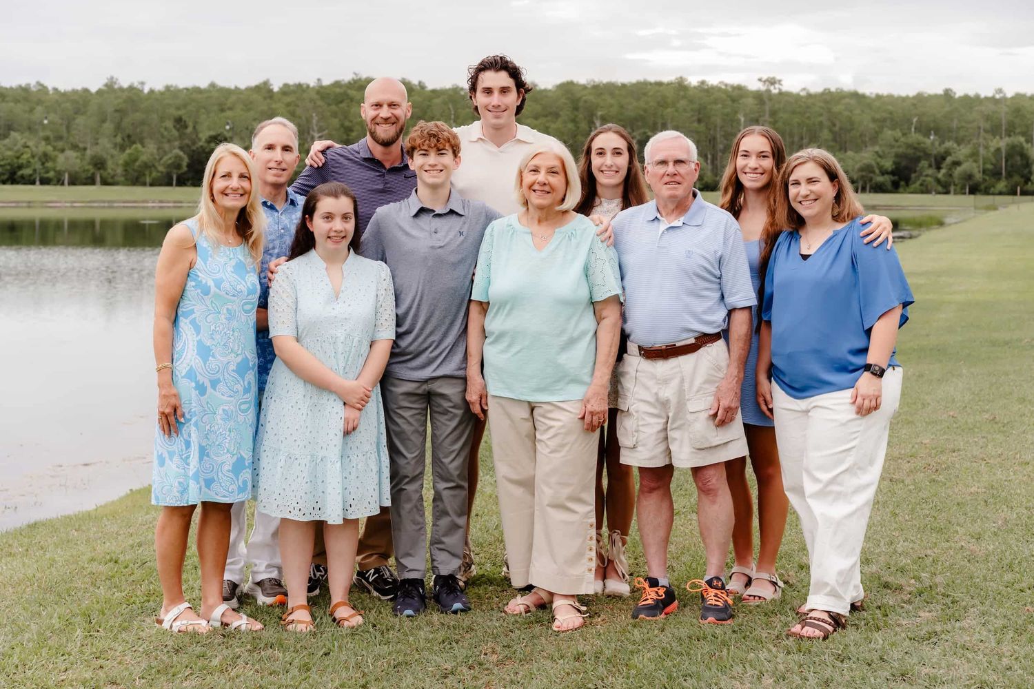 Large family group portrait smiling outdoors near a lake, dressed in coordinating blue and neutral tones.
