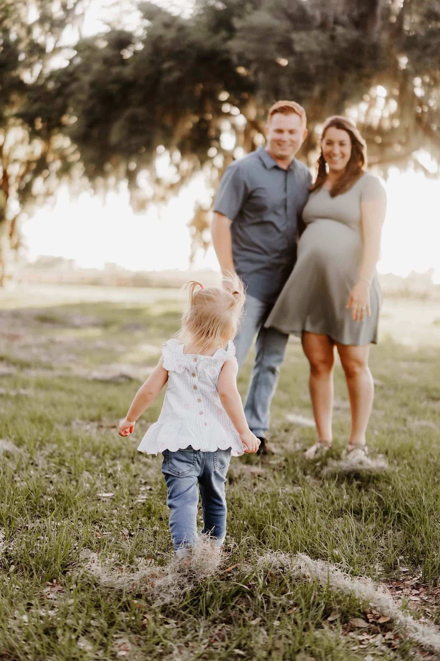 A pregnant woman and man stand in a sunny park while a toddler girl in jeans walks toward them.
