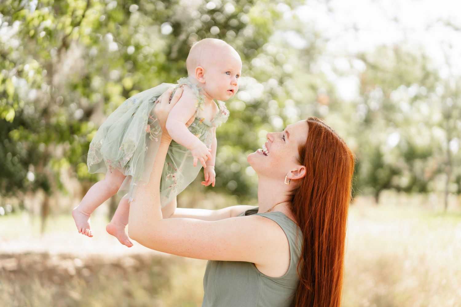 A parent lifts and plays with their baby in a sunny outdoor field during a joyful bonding moment.
