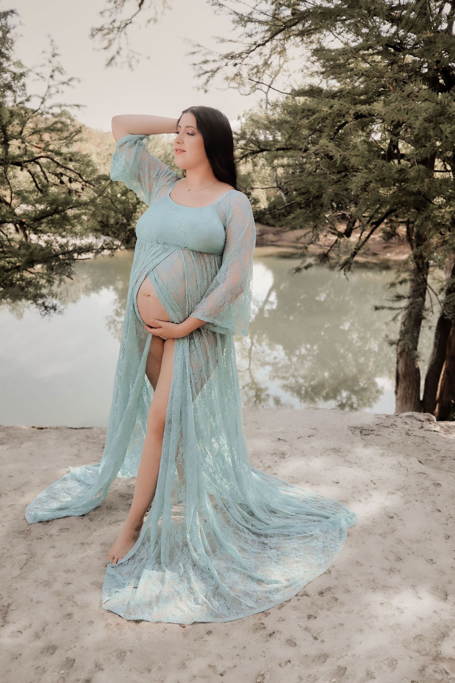 Pregnant woman in flowing teal lace maternity gown poses gracefully by a serene lake surrounded by trees.