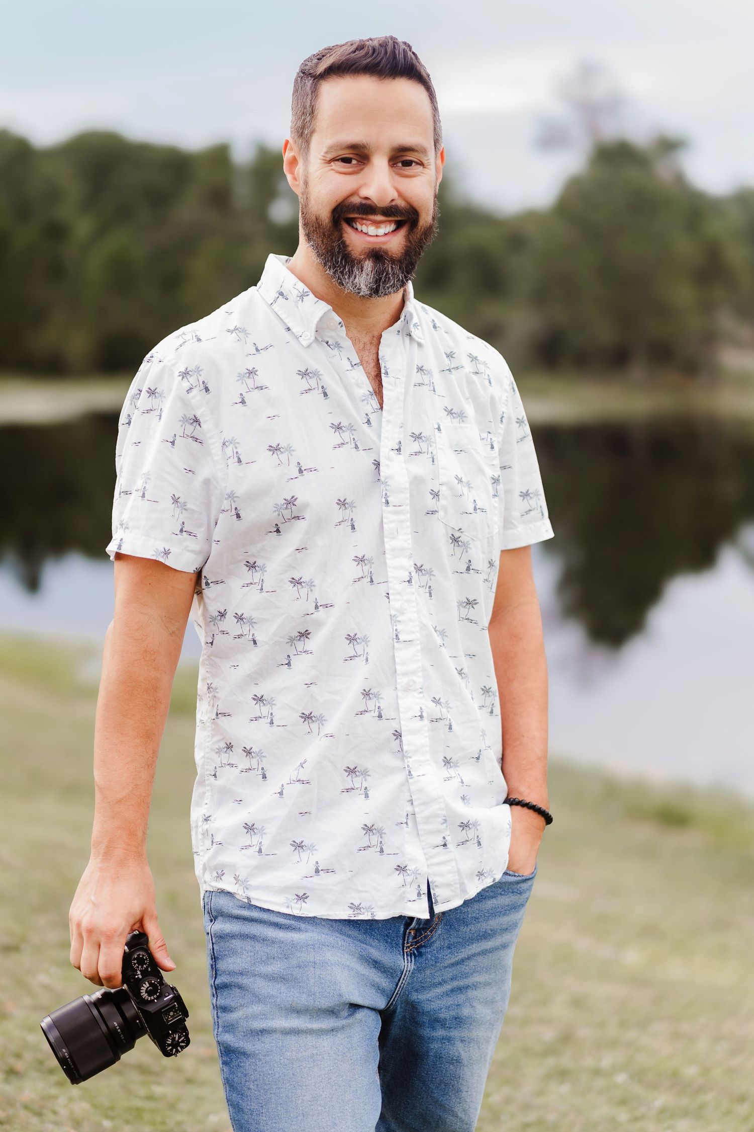 Smiling bearded man holding a camera wearing a white palm tree print shirt and jeans standing near a lake.