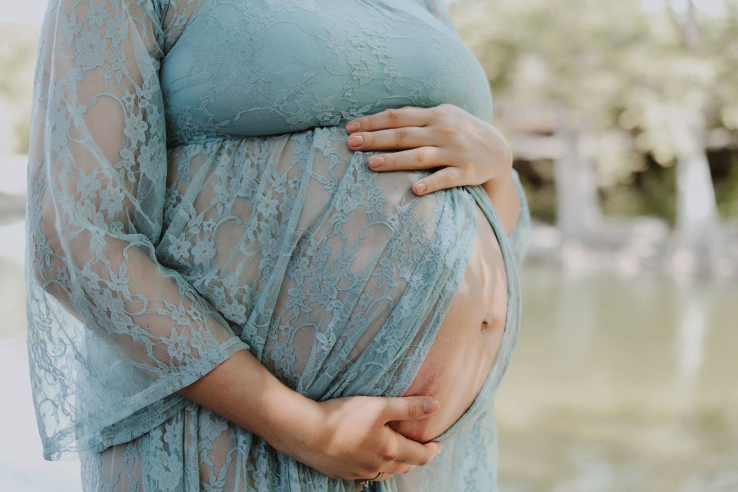 Pregnant woman in teal lace dress cradling her bare baby bump outdoors near water.