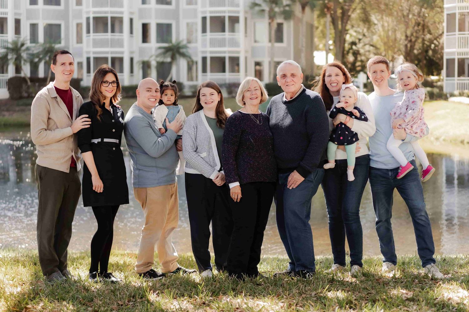 Large multigenerational family posing outdoors near a pond with resort buildings in the background.