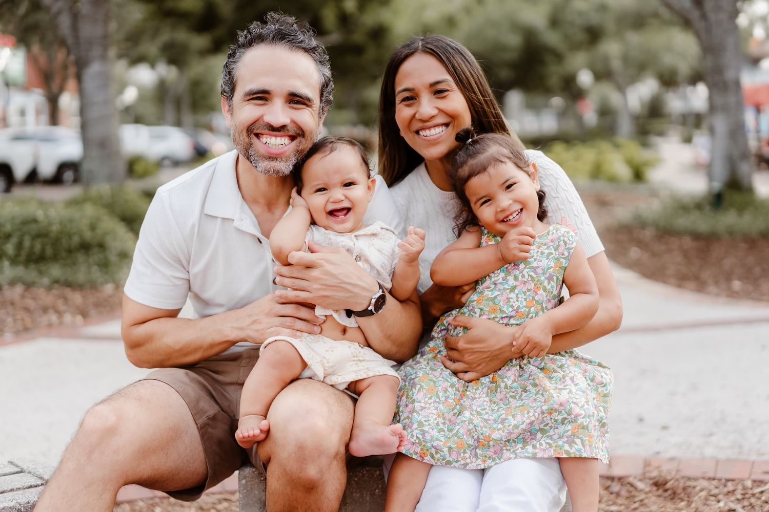 Happy family of four smiling outdoors, parents holding two young children in a park setting.