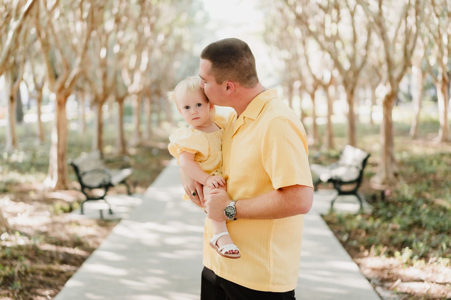 A man in a yellow shirt kisses a baby in a yellow outfit while standing on a park path surrounded by trees.