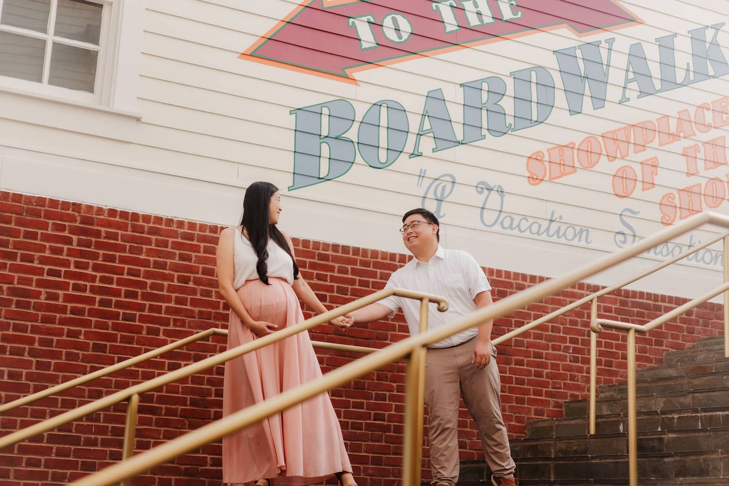 Couple standing on golden stairs by a colorful Boardwalk sign on a brick wall, woman in pink dress, man in white shirt.