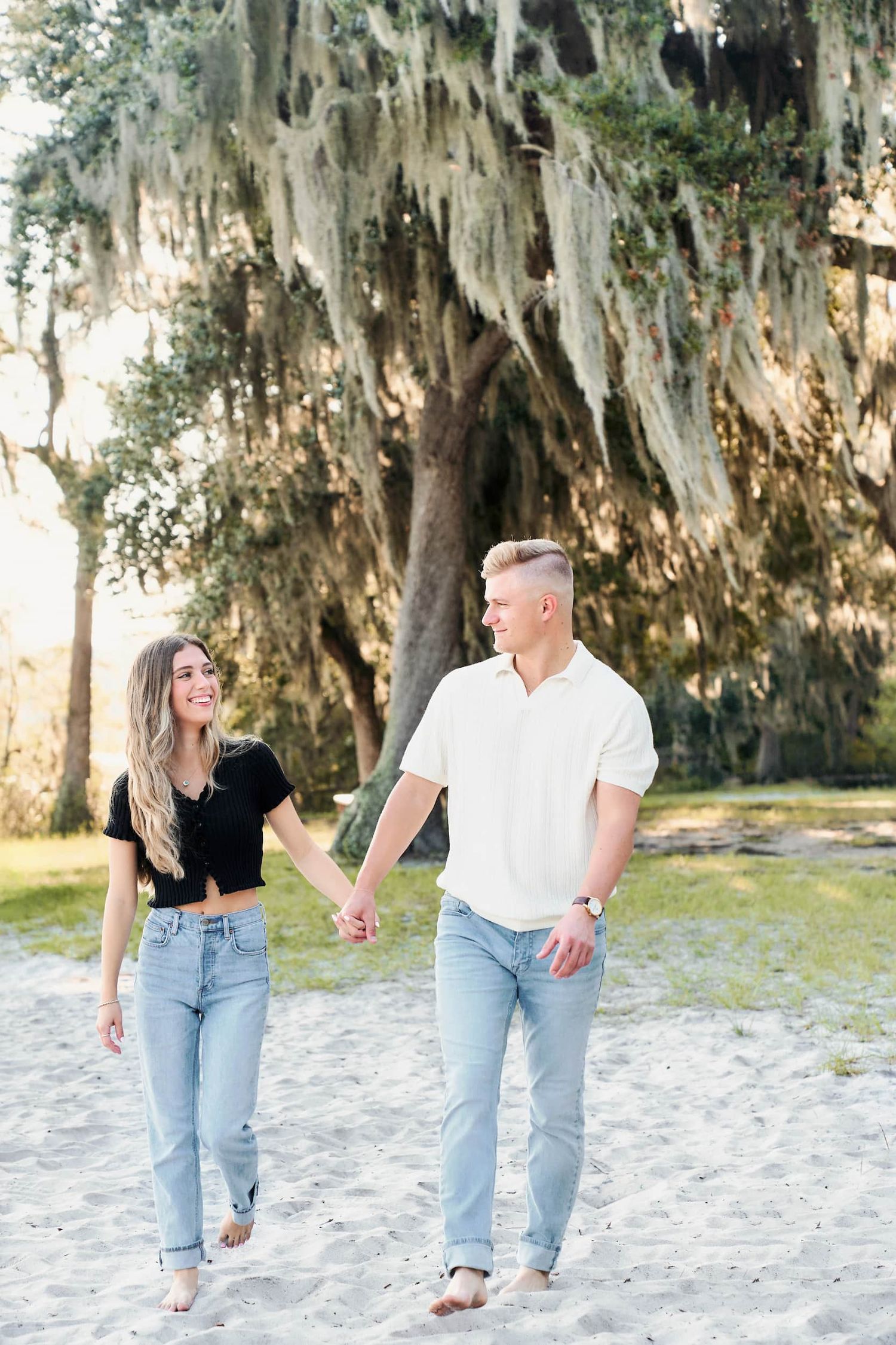 A couple holds hands and walks barefoot on sandy ground beneath moss-draped trees in warm golden light.