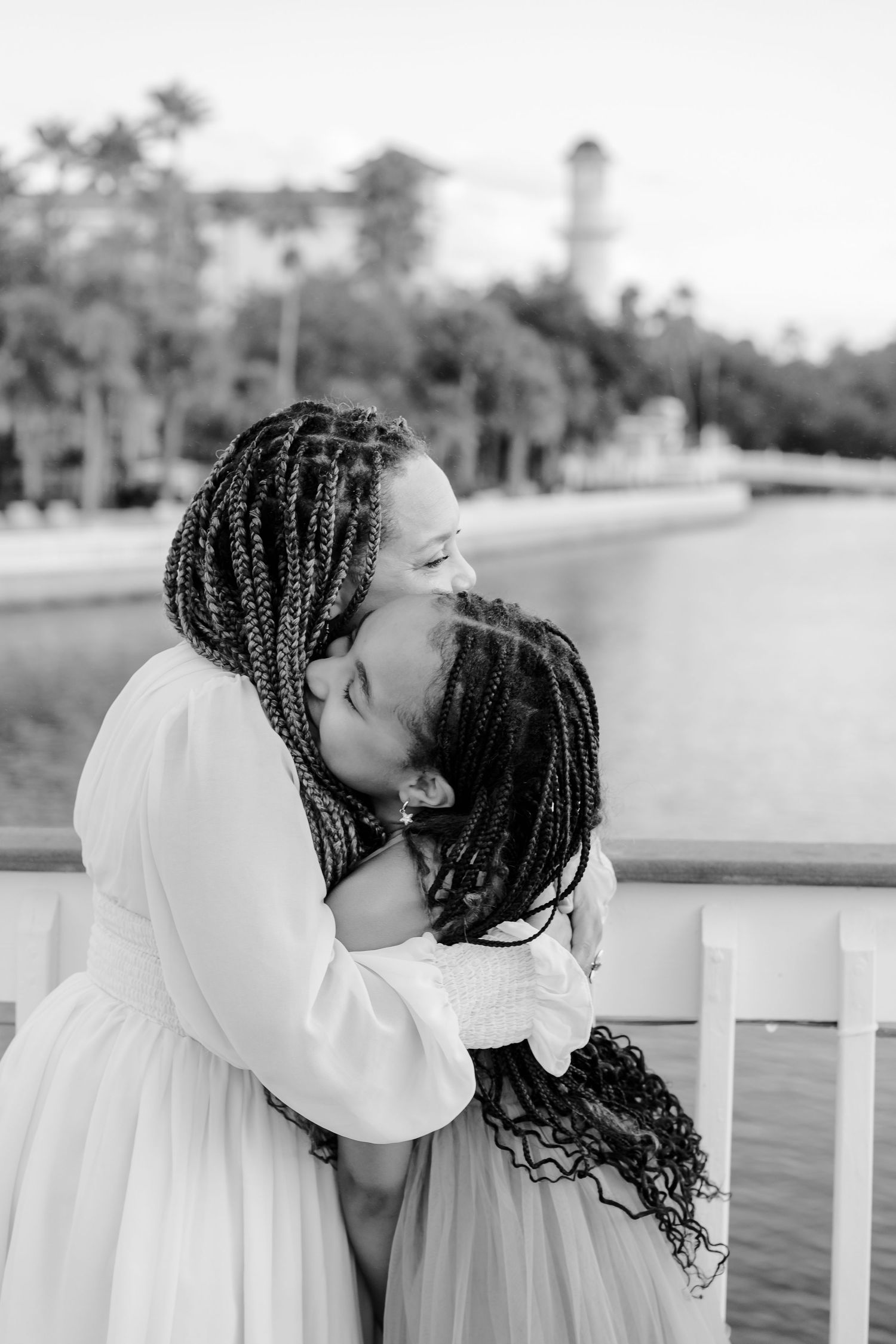 Two women with long braided hair share a tender embrace on a waterfront pier, wearing flowing light dresses in a black and white photo.
