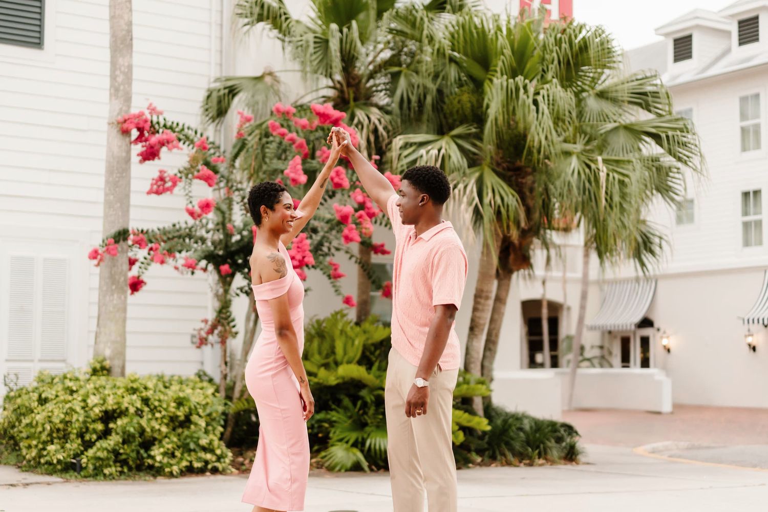 Couple in pink outfits dancing outdoors near palm trees and bougainvillea by a white building.