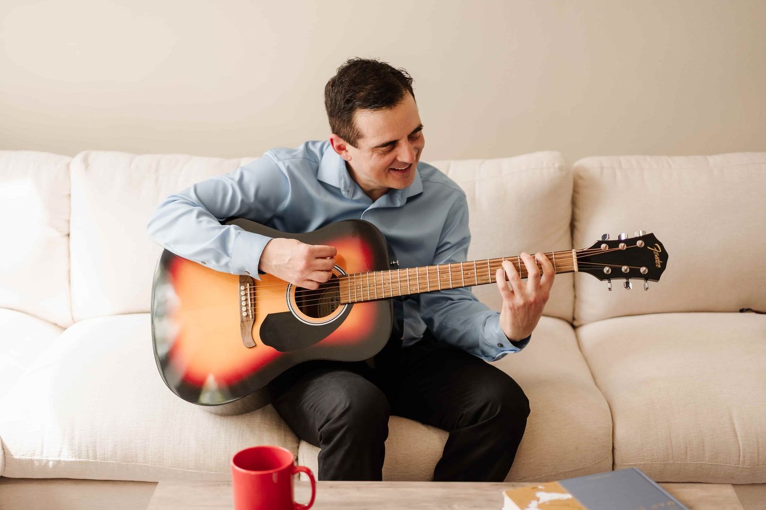 Man playing acoustic guitar on white sofa with red mug on coffee table in bright living room.