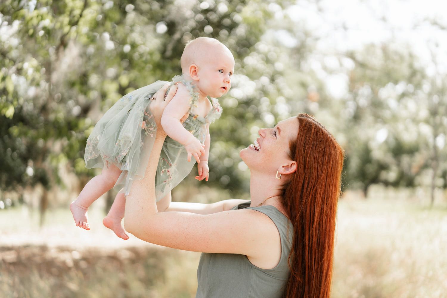 A joyful redhead mother lifts her baby girl in a green dress outdoors in a sunlit park setting.