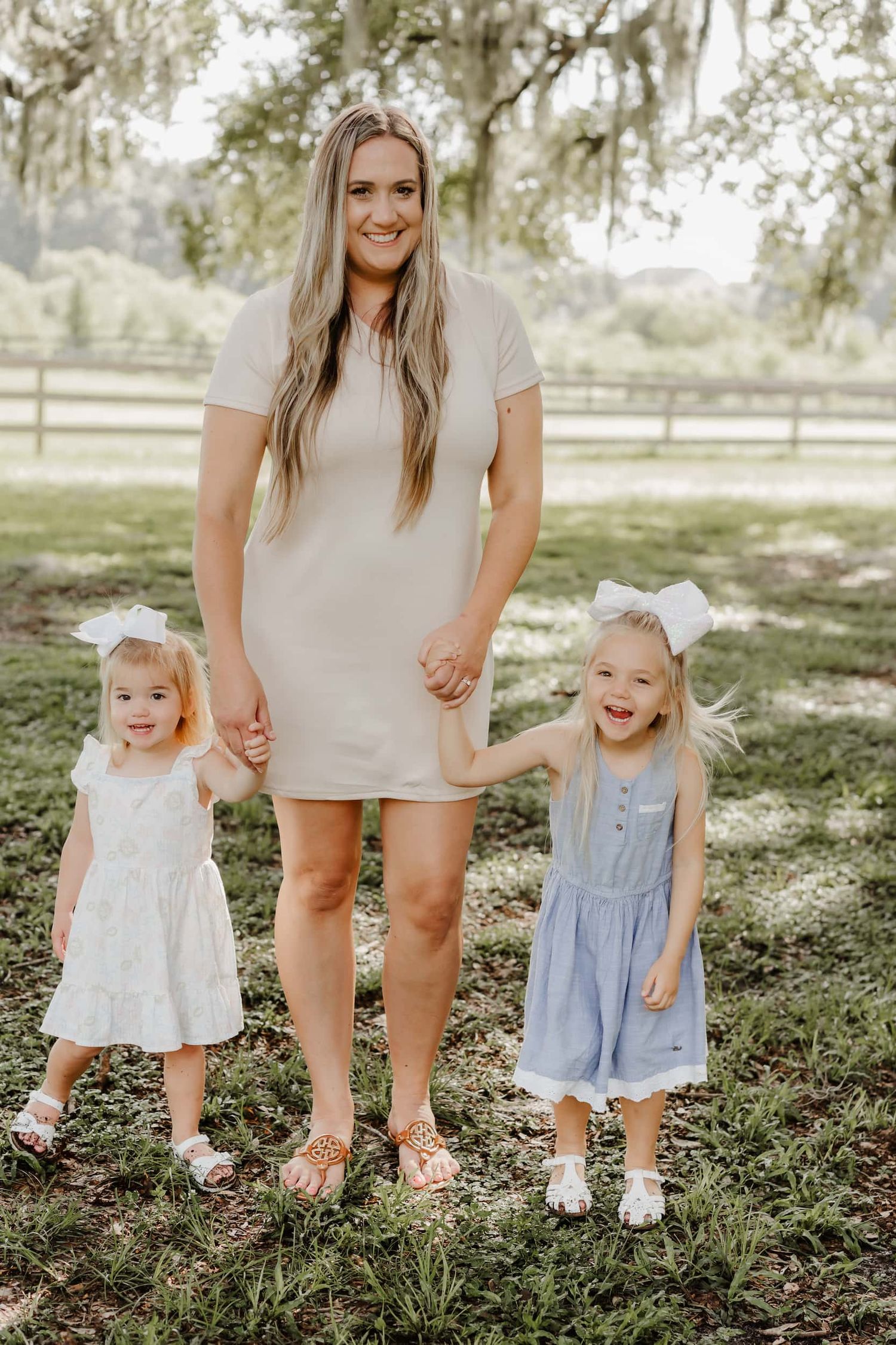 A smiling woman in a beige dress holds hands with two young girls in white and blue dresses outdoors.