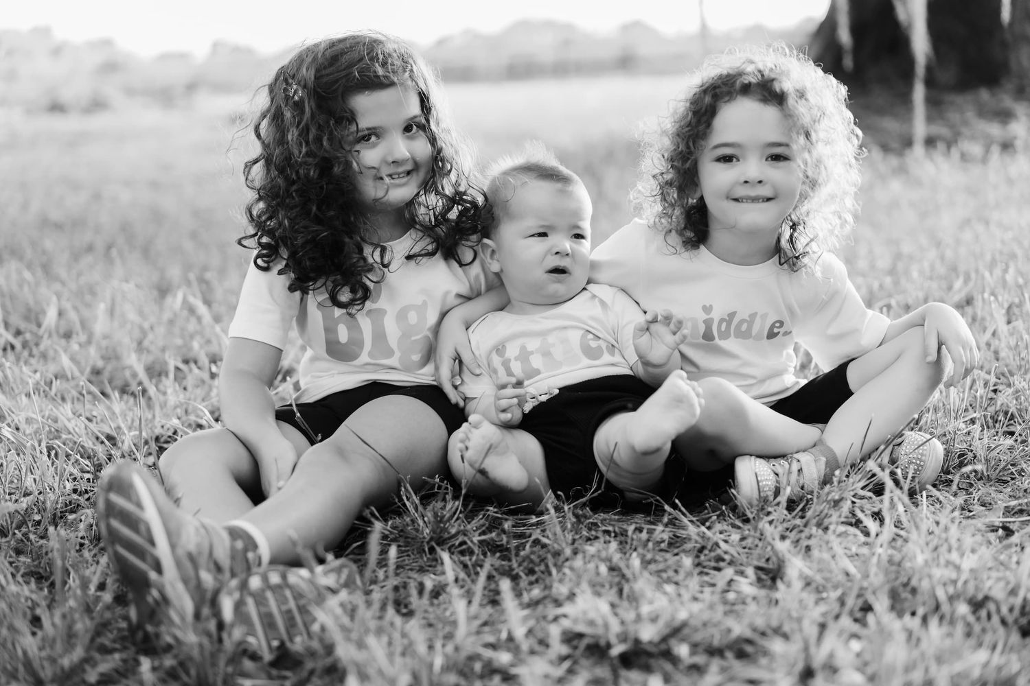 Black and white photo of three young siblings sitting together on grass, two girls with curly hair and a baby between them.