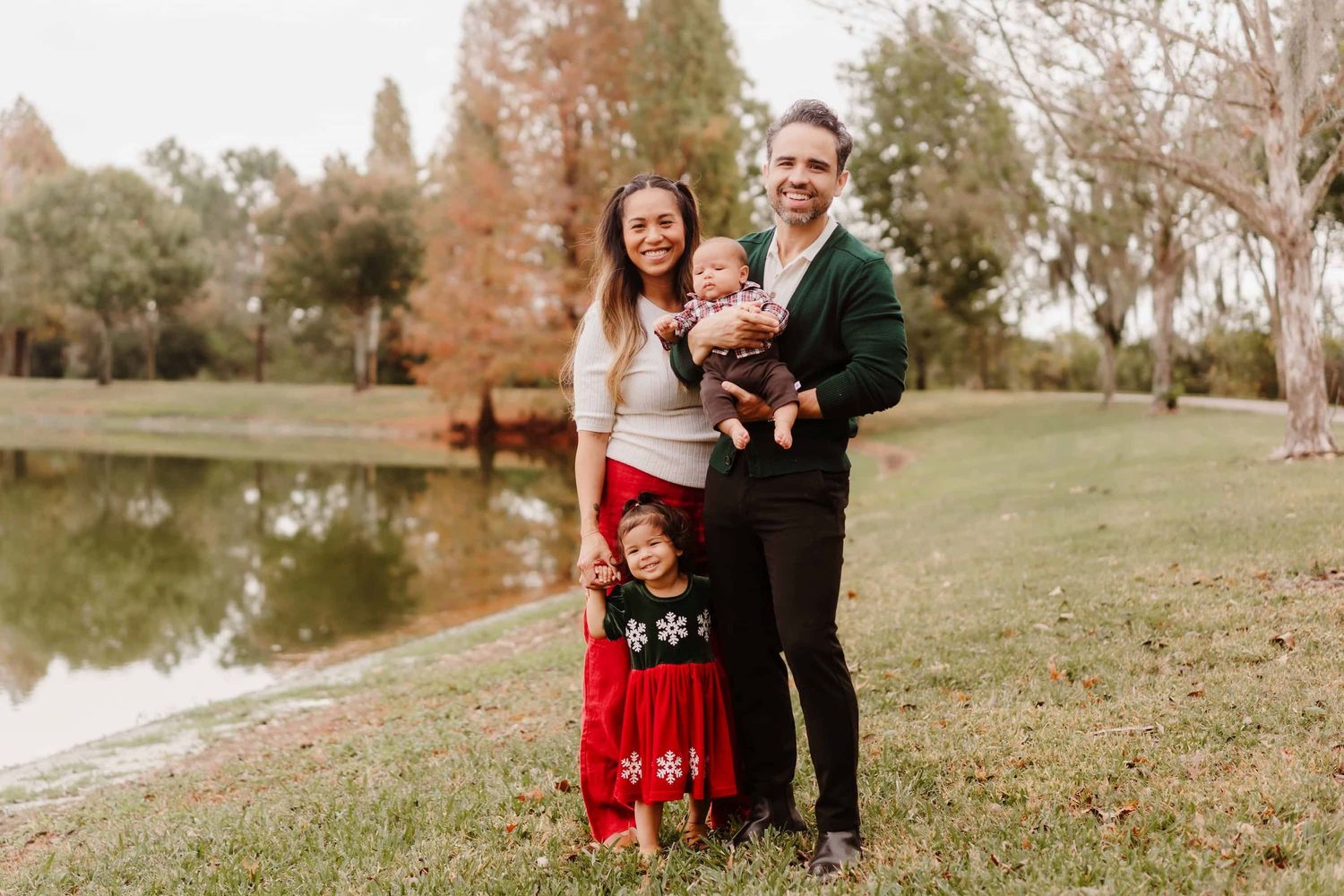 Family of four posing outdoors near a pond in festive holiday attire, smiling together on a grassy lawn.