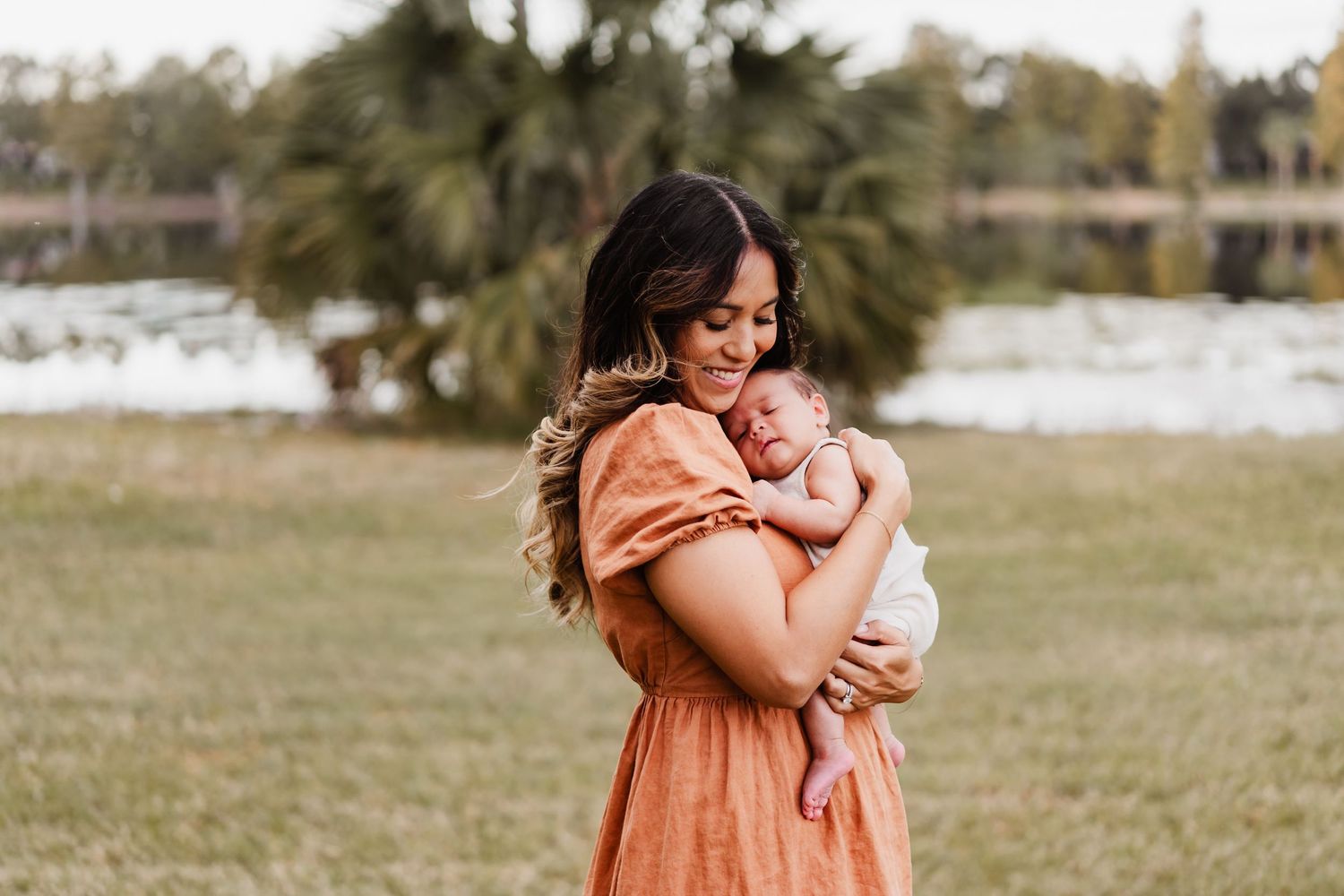 A couple embraces tenderly in a grassy field on a sunny day wearing casual earth-toned clothing.