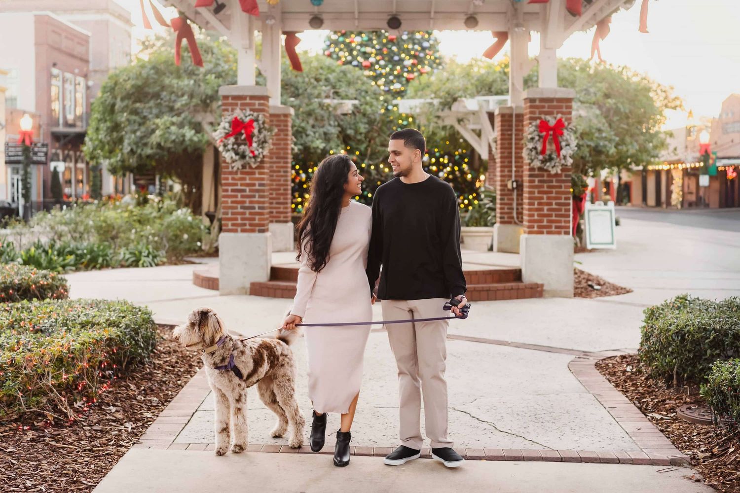 Couple walking dog at Christmas decorated outdoor shopping center with brick pillars and holiday wreaths.