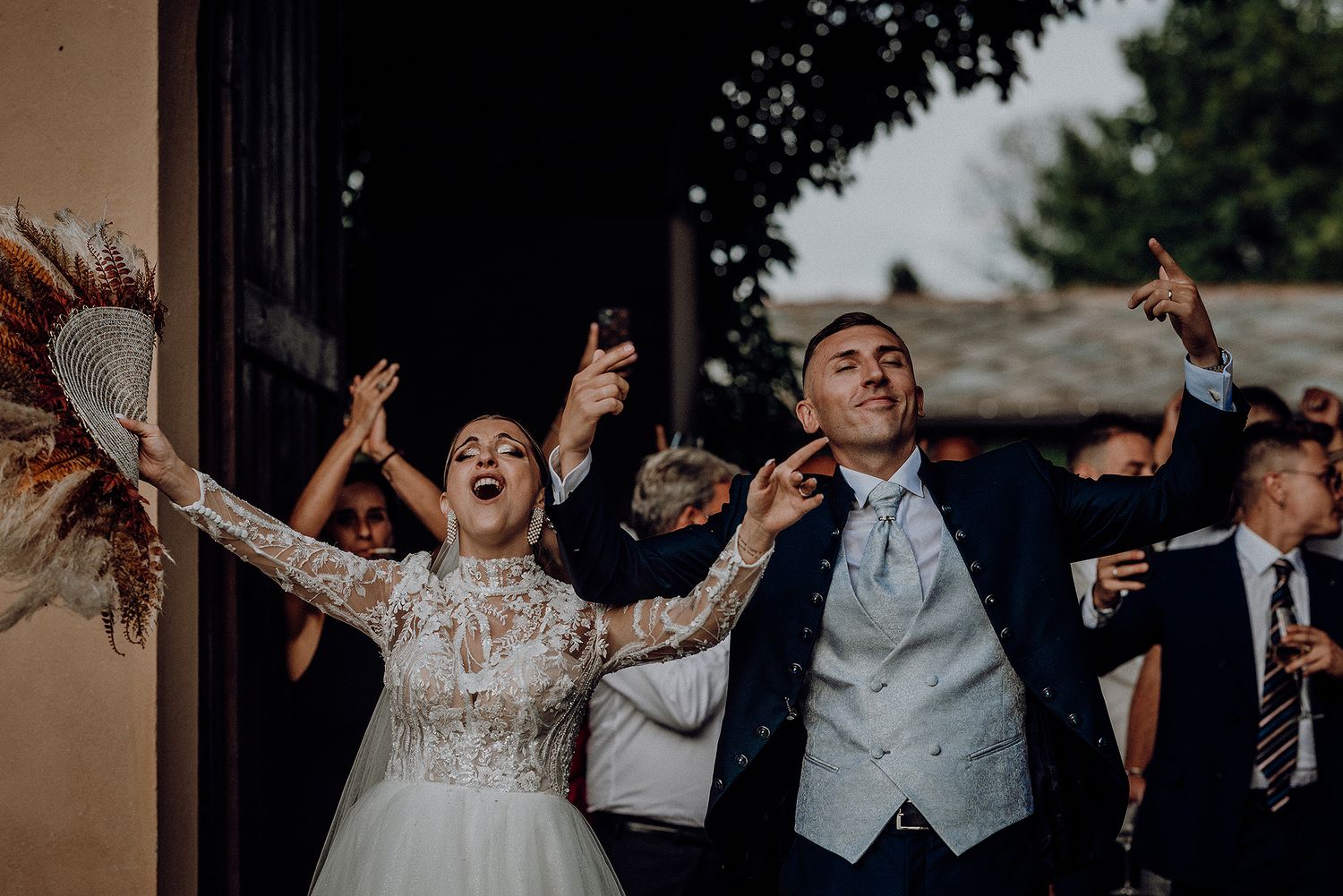 Excited bride and groom celebrate with arms raised at wedding venue entrance, guests cheering in background.