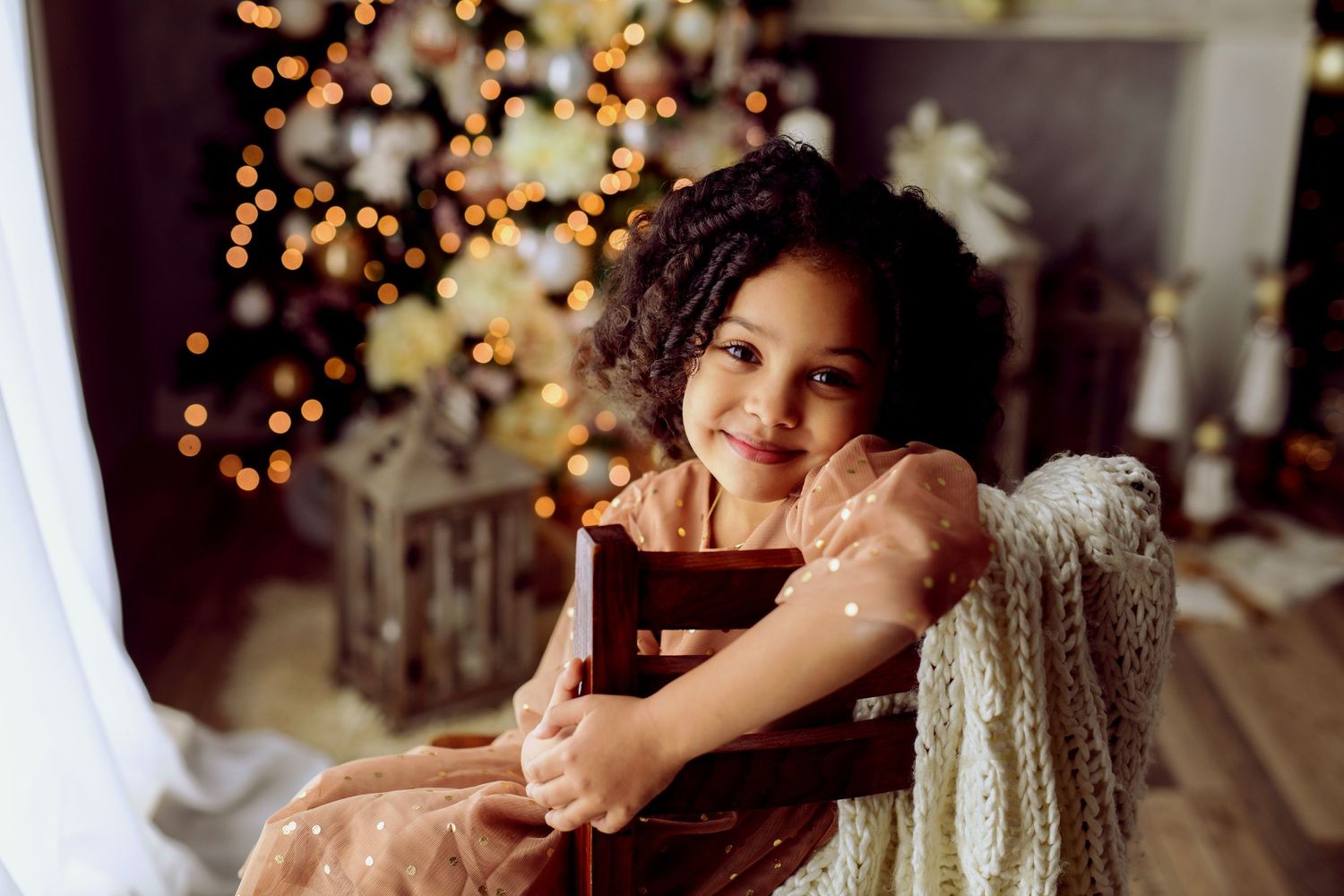 Happy curly-haired girl in sparkly dress sitting on chair near glowing Christmas tree.