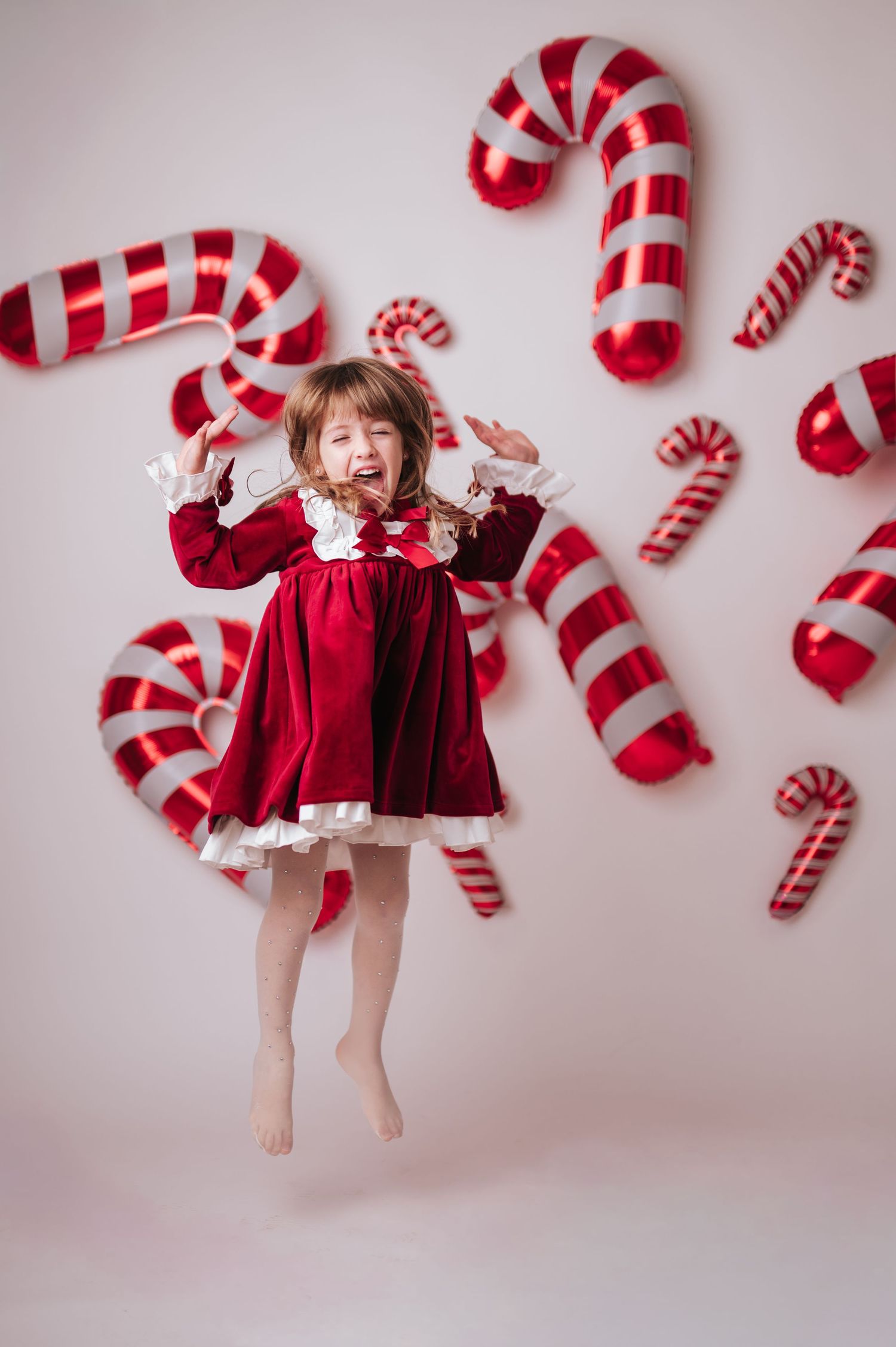 Excited little girl in red dress jumping among large candy cane balloons on white background.