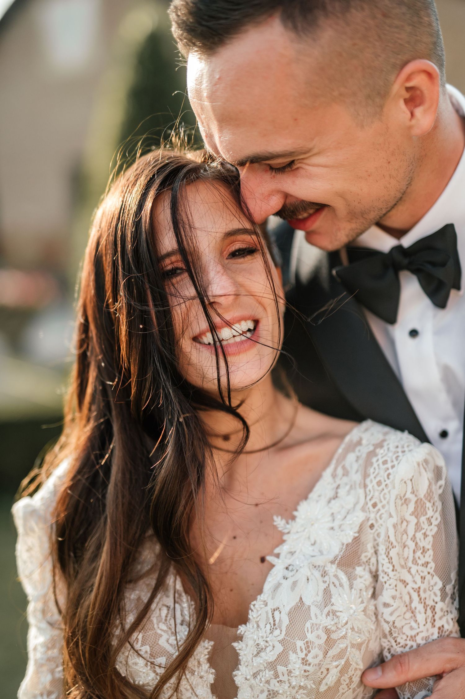 Radiant bride with windswept hair laughing joyfully with smiling groom in bow tie at wedding.